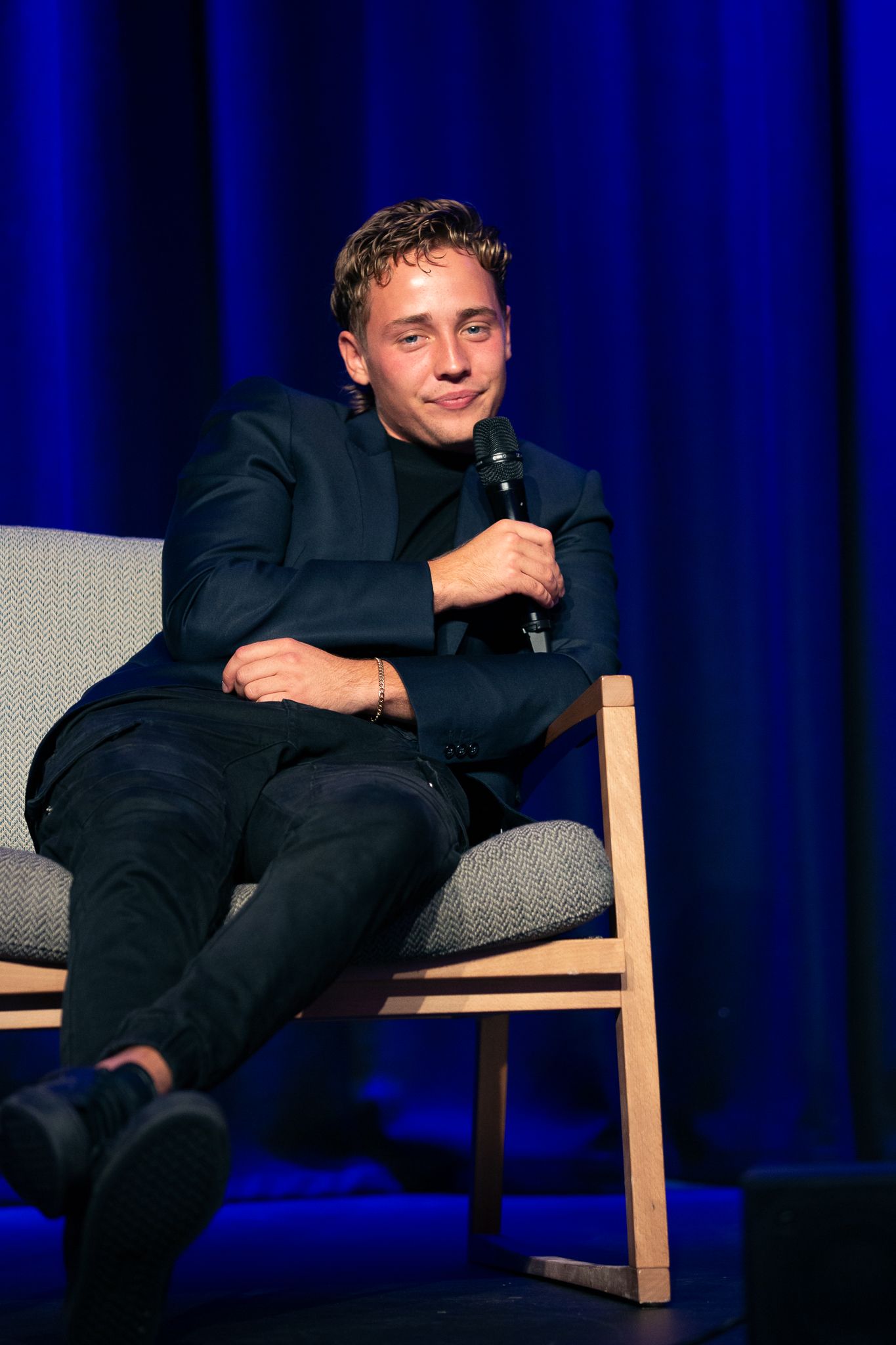 Person in dark clothing seated on stage during presentation with blue curtain backdrop.