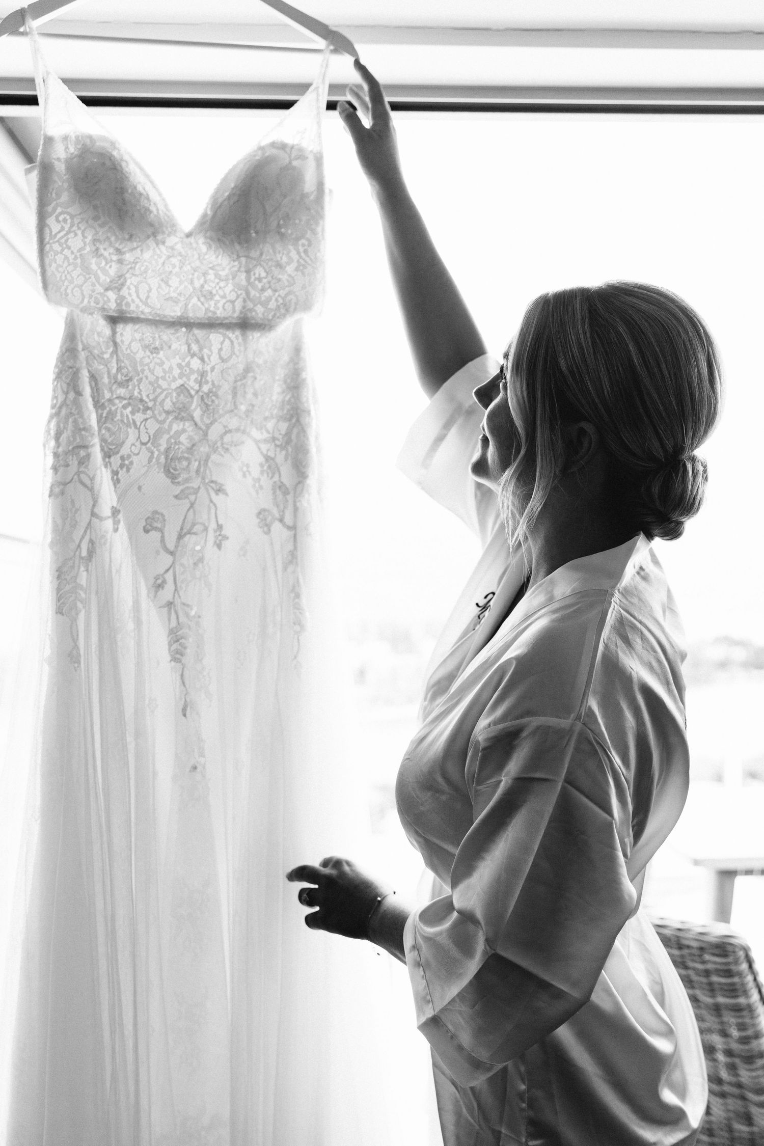 Woman in white shirt looking up at hanging wedding dress in black and white photo.