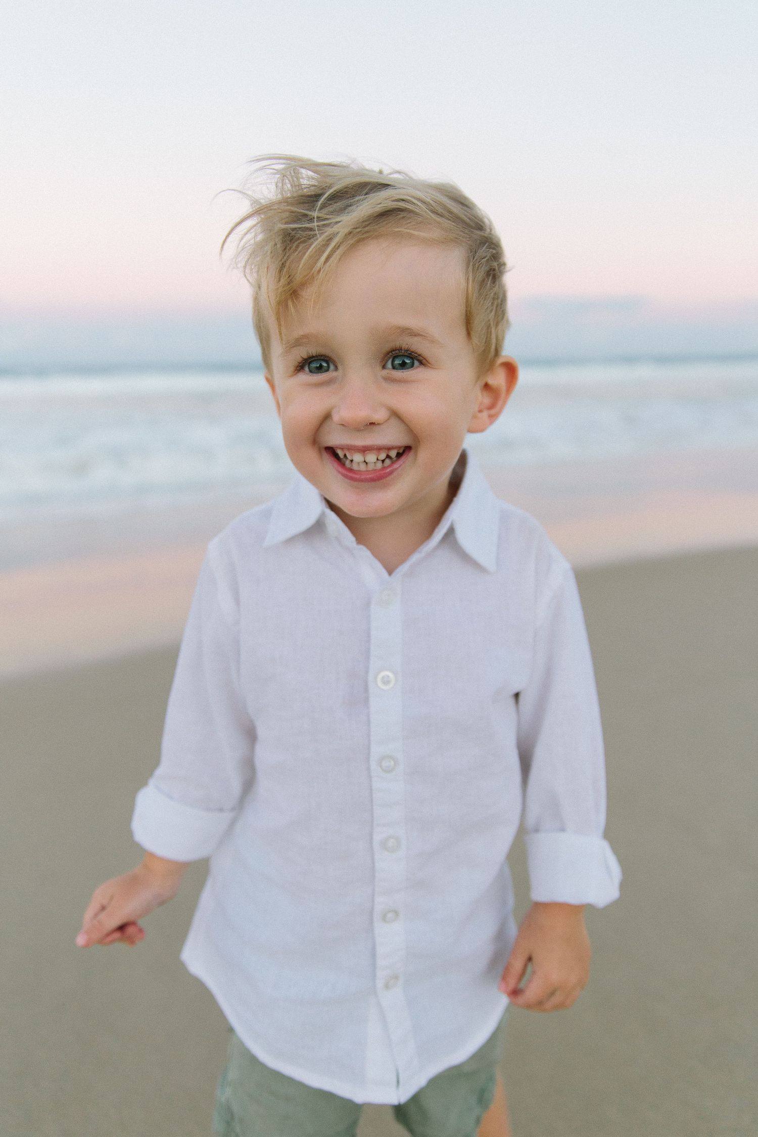 Child in white button-down shirt smiling and playing on the beach at sunset.