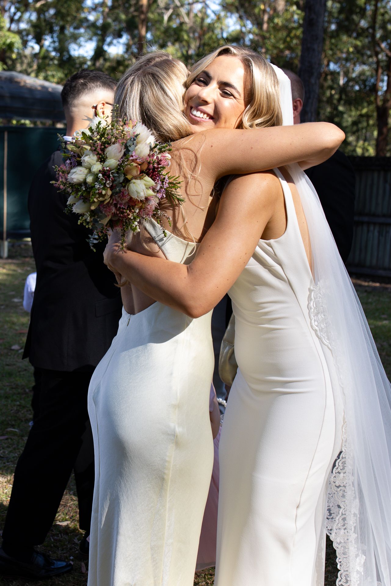 Two women in white dresses share a joyful embrace at an outdoor wedding celebration.
