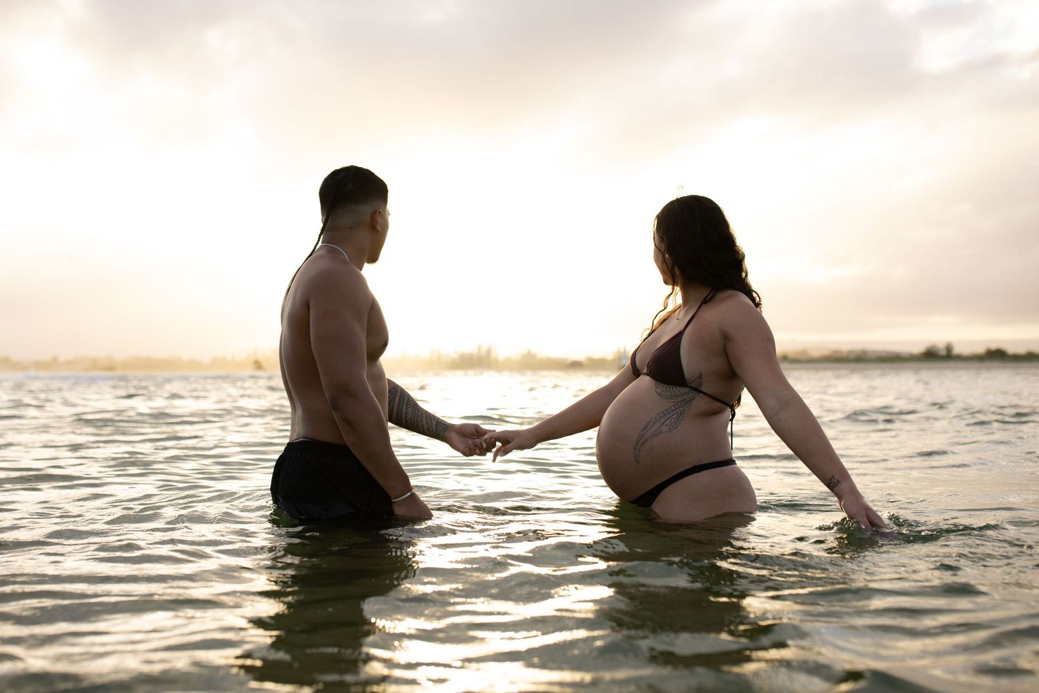 Couple holding hands in ocean water during sunset maternity photoshoot.