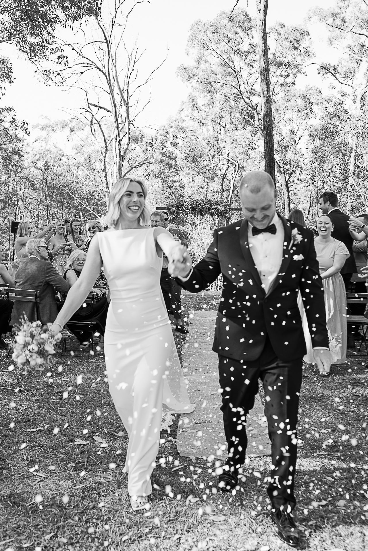 Black and white photo of wedding party walking through falling confetti outdoors.