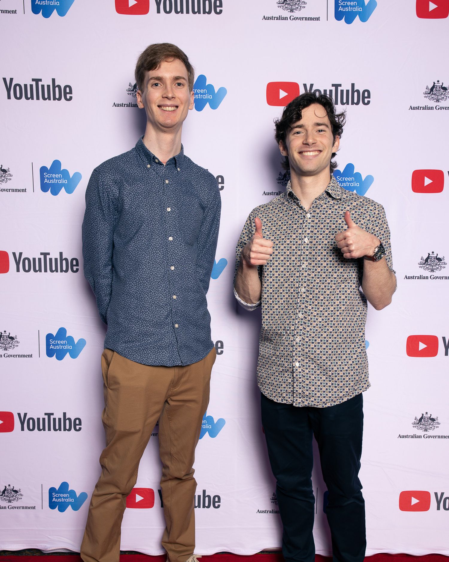 Two people posing together at a YouTube event with branded backdrop and logos.