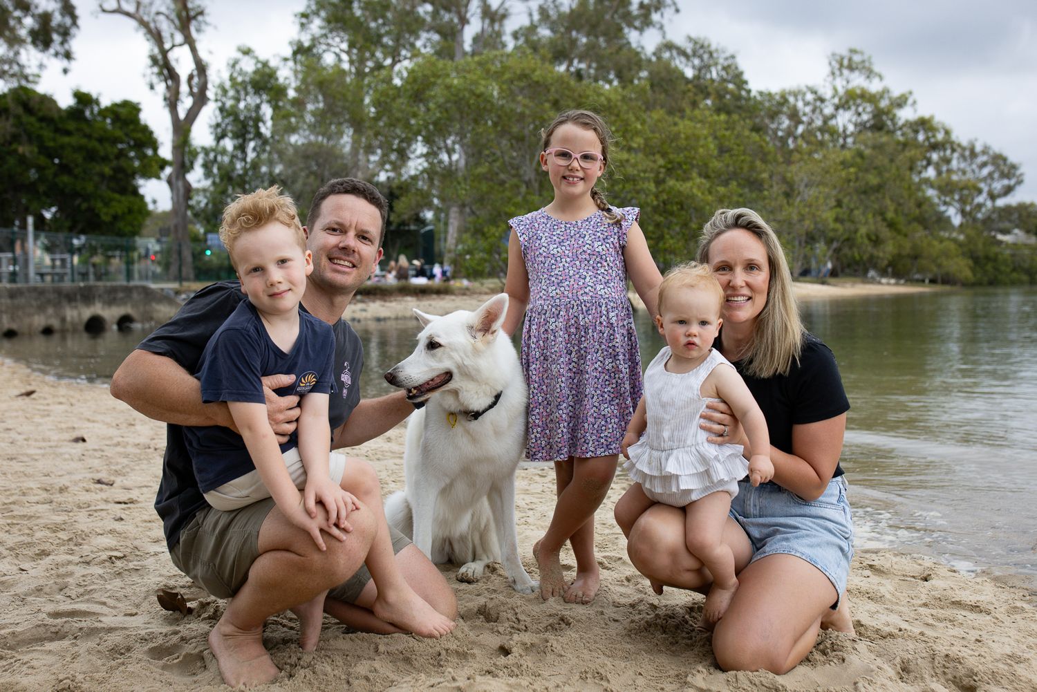 A family and their white dog posing together on a sandy beach near water.