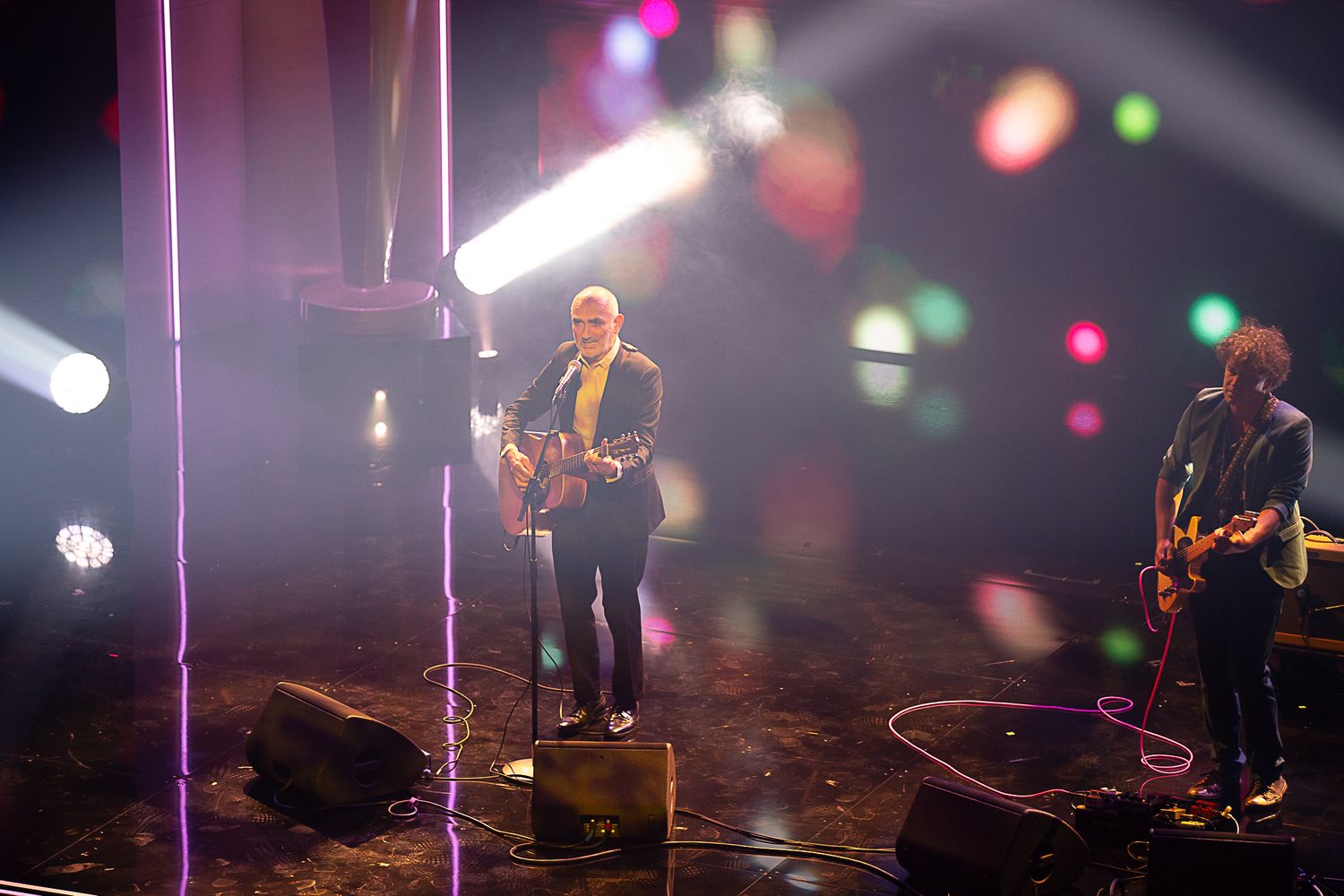 Concert stage with atmospheric lighting and performers silhouetted against colorful spotlights.