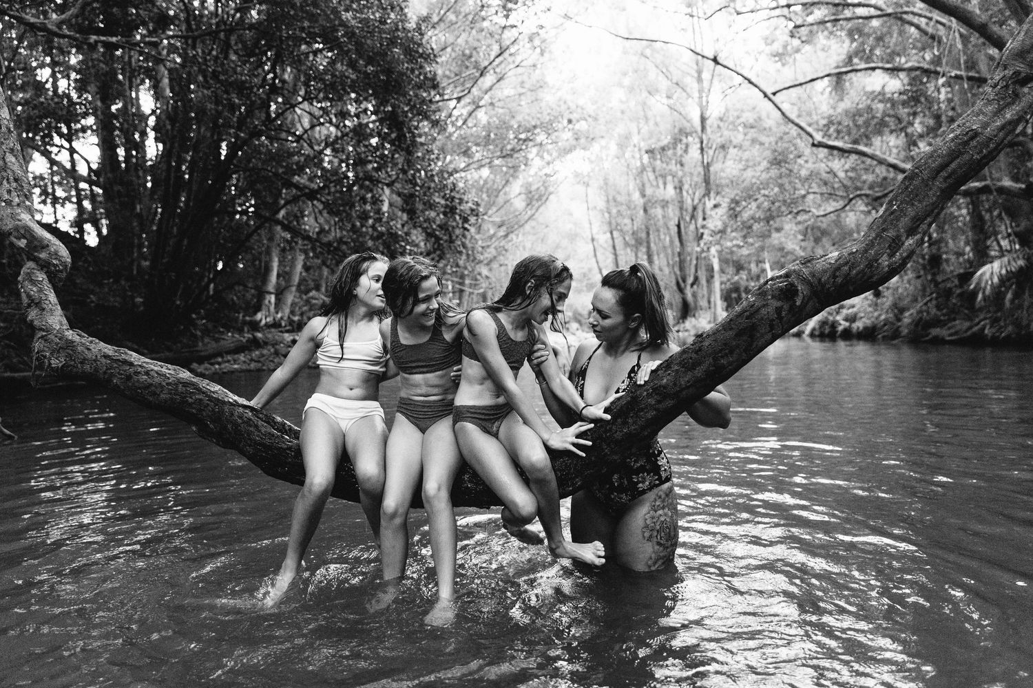 Group of people sitting and playing on a fallen tree over water in a natural setting.