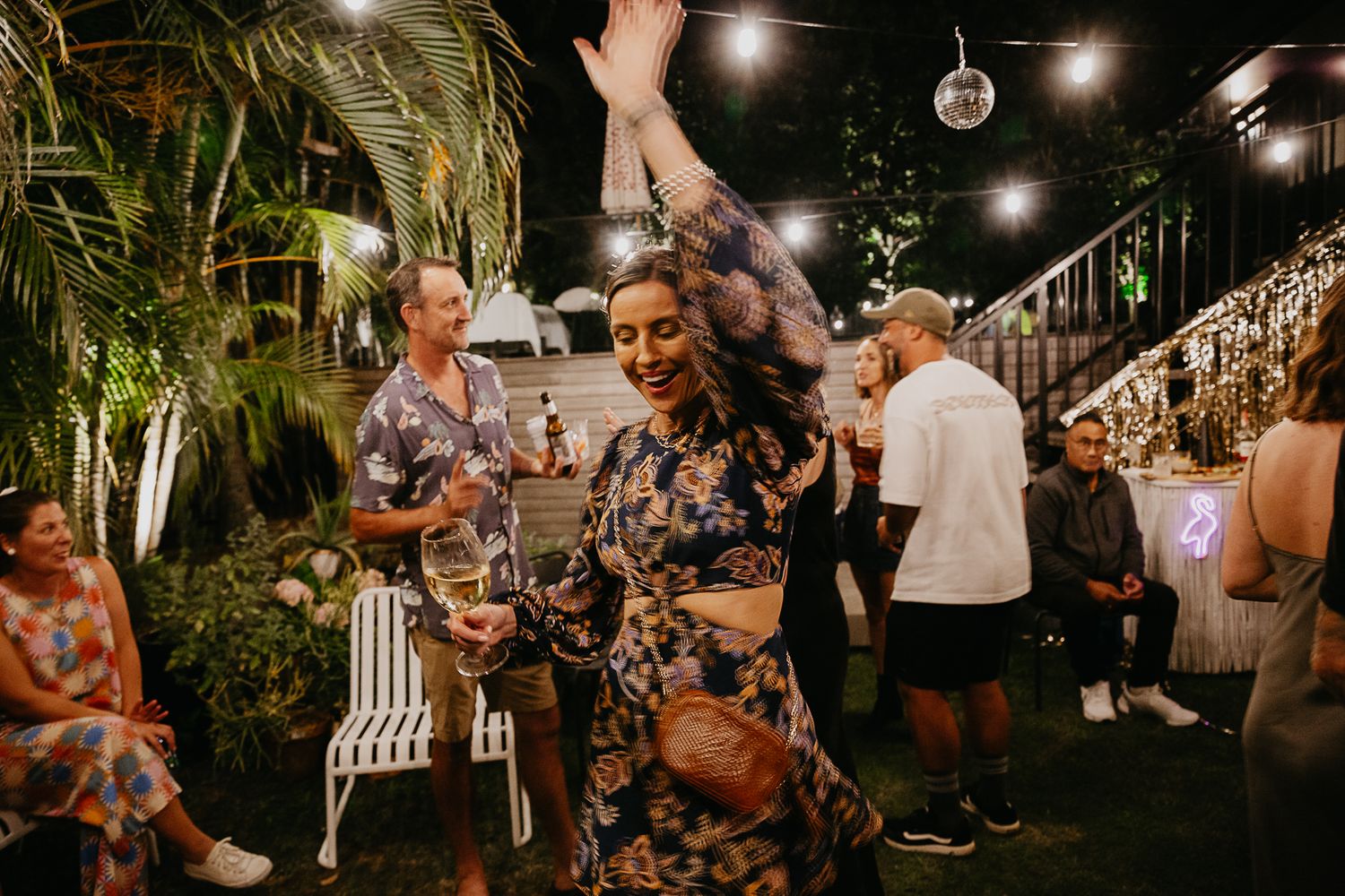 Nighttime outdoor wedding reception with guests dancing under string lights and palm trees.