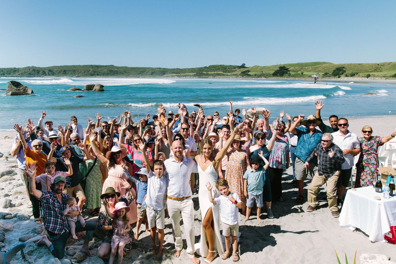 Large group photo of wedding guests gathered on a sunny beach with ocean views.