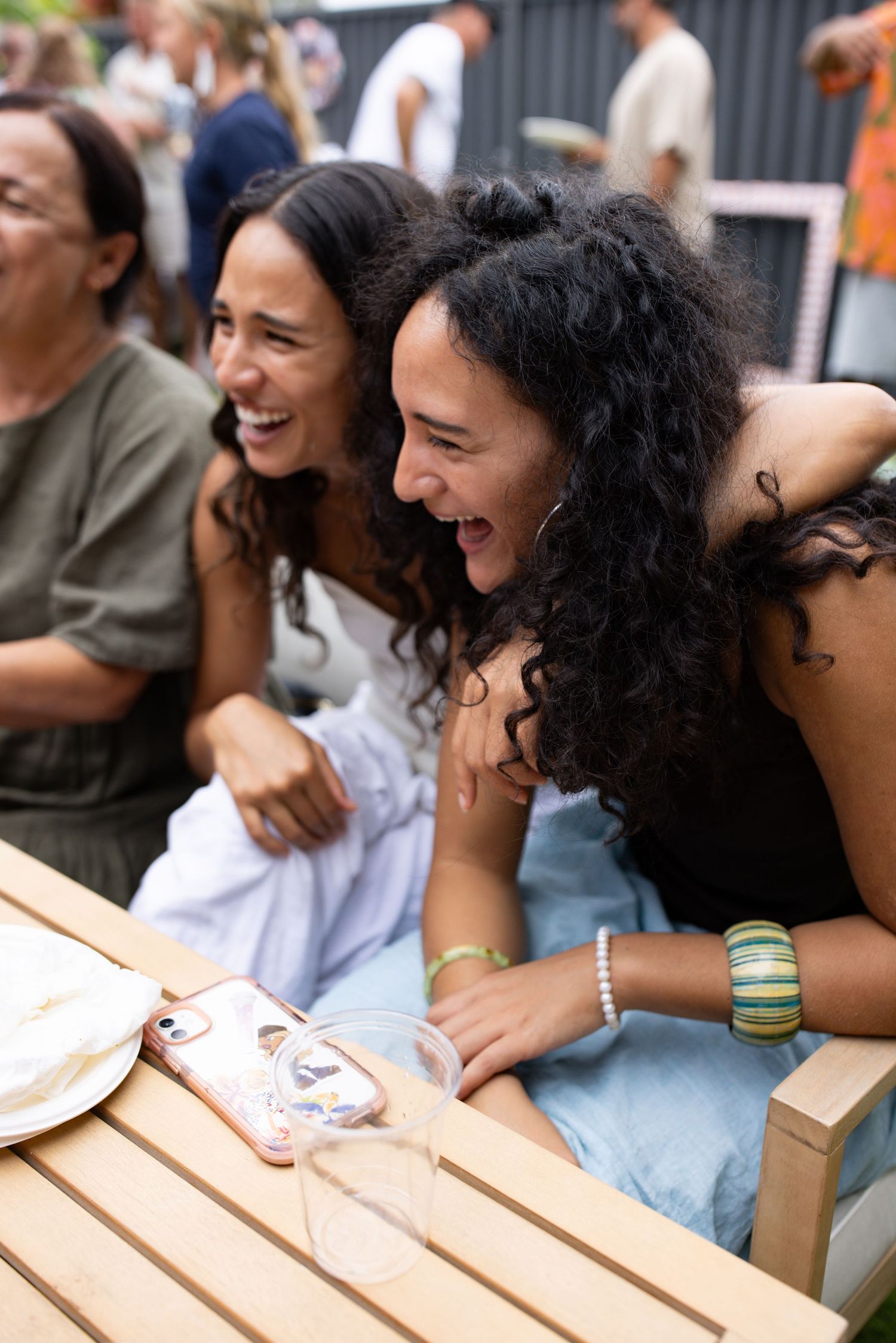 Friends sharing laughter while sitting at an outdoor wooden table during a gathering.