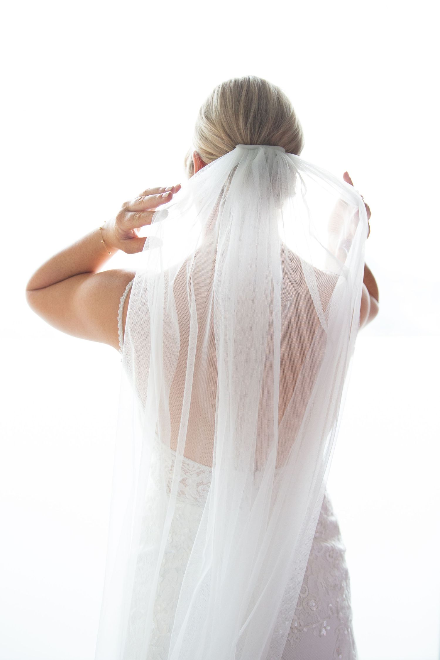 Back view of bride adjusting her flowing white veil against bright background.