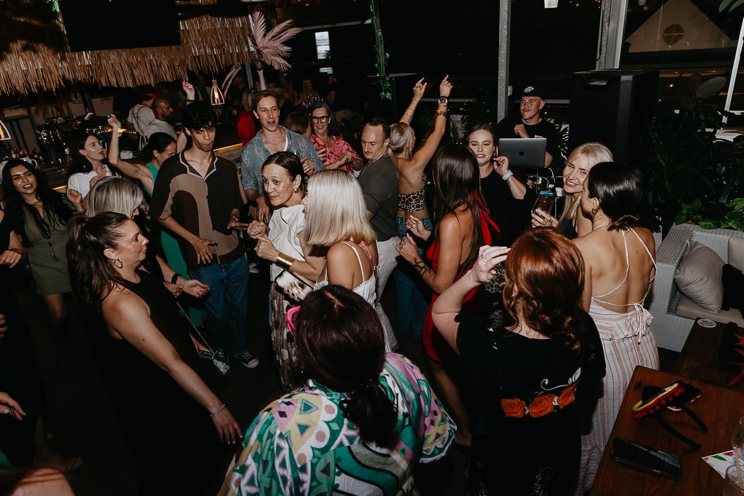 Crowded dance floor at a nighttime wedding reception with tropical decor.