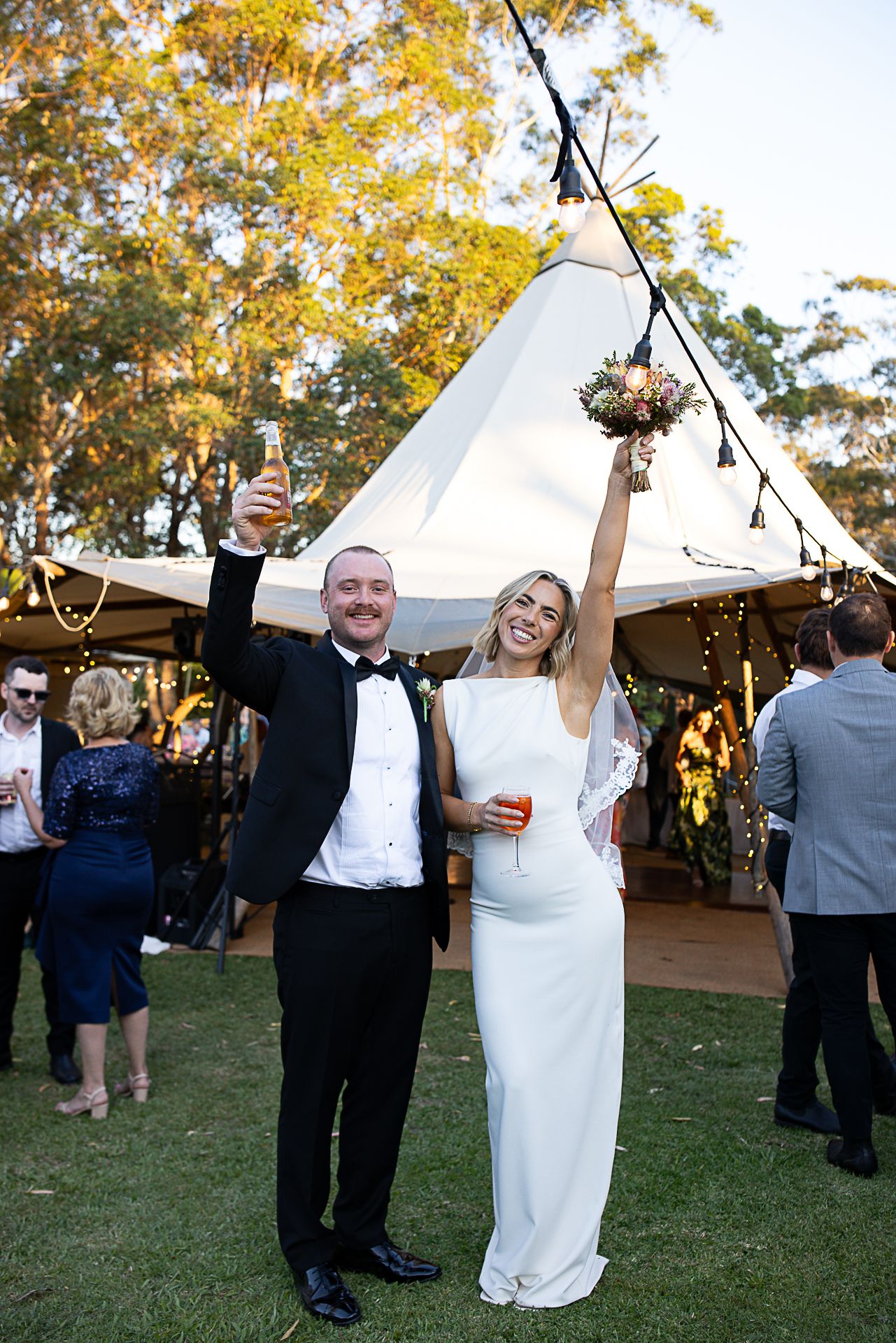 Newlyweds celebrate joyfully on grass lawn with reception tent in background.