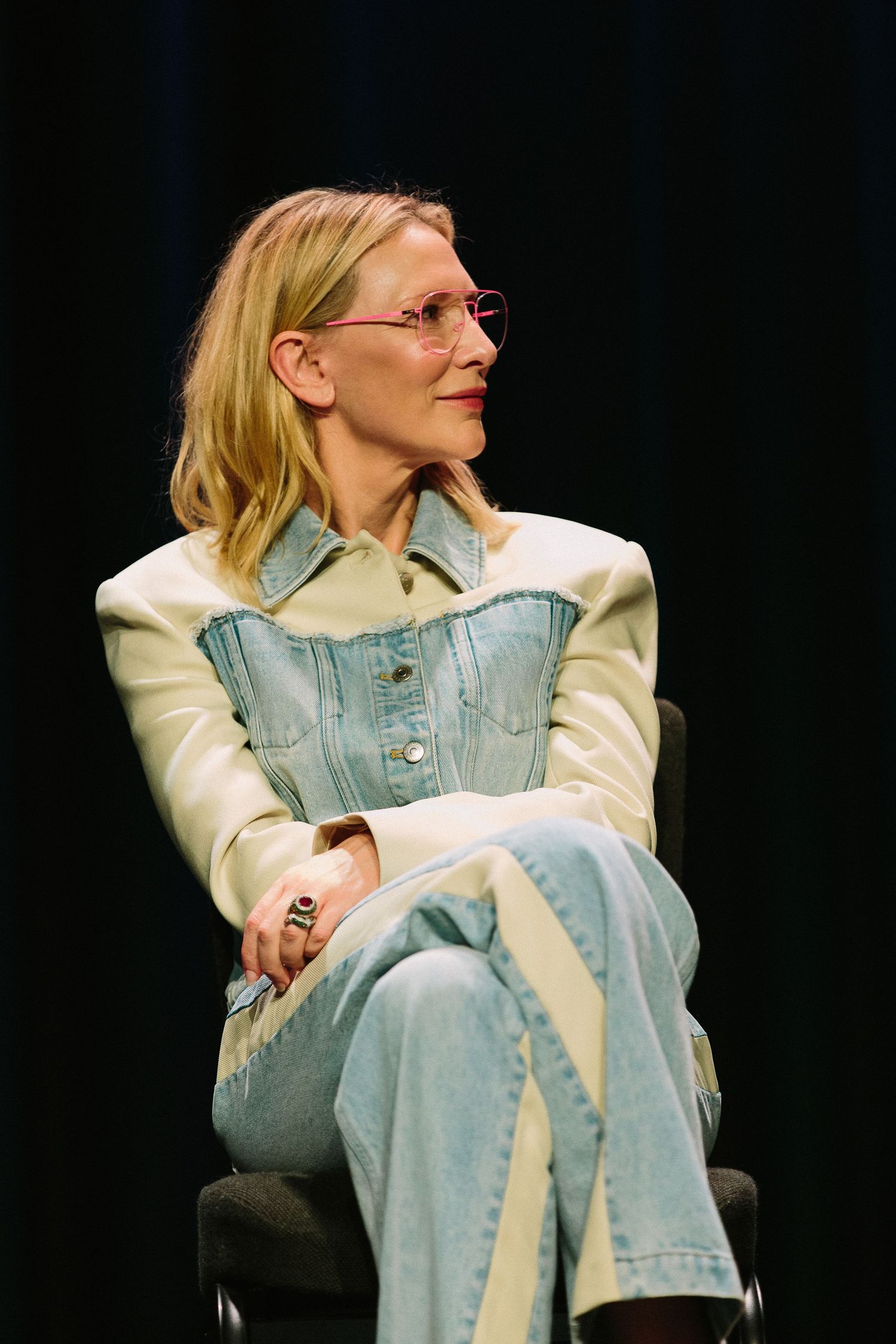 Person in denim jacket and white sleeves sitting thoughtfully against dark background.