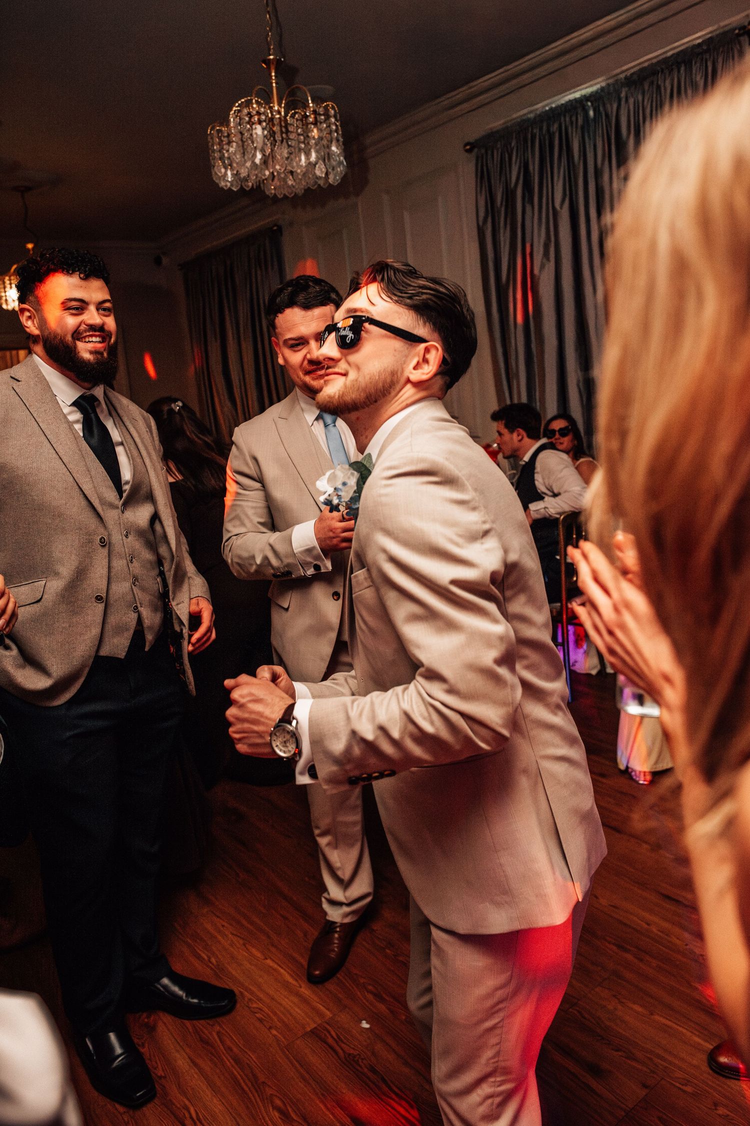 Groomsmen in gray suits dancing and celebrating at a wedding reception under a chandelier.