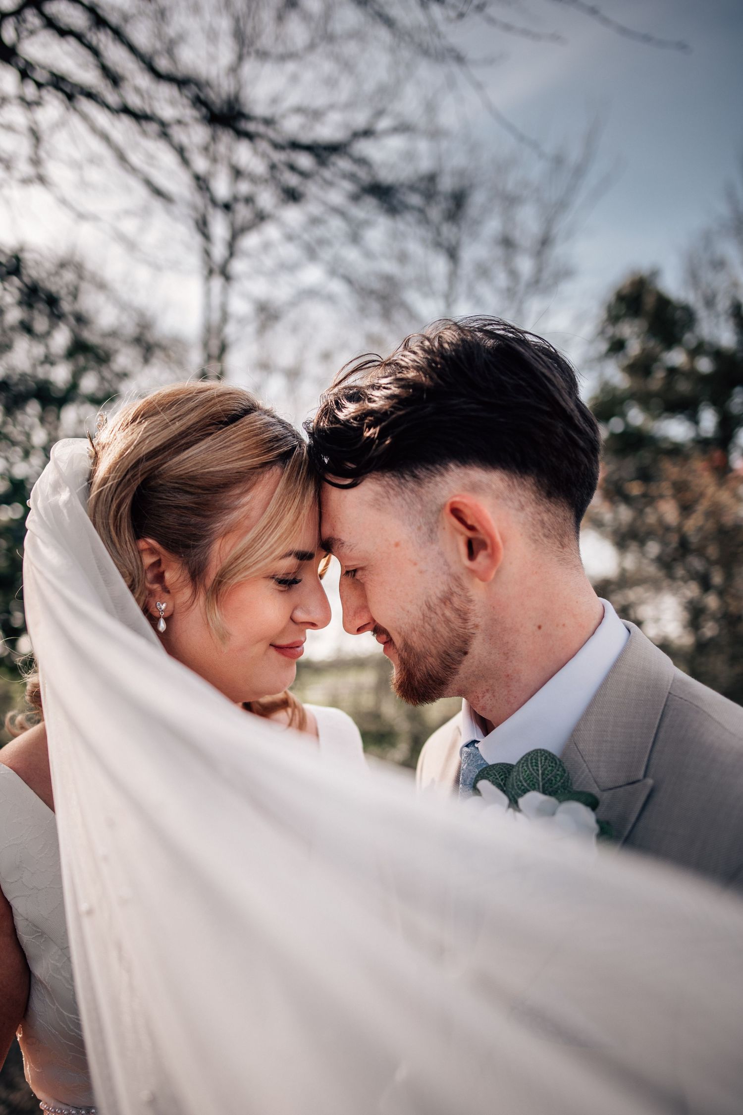 Bride and groom touching foreheads romantically under a flowing white veil outdoors on their wedding day.