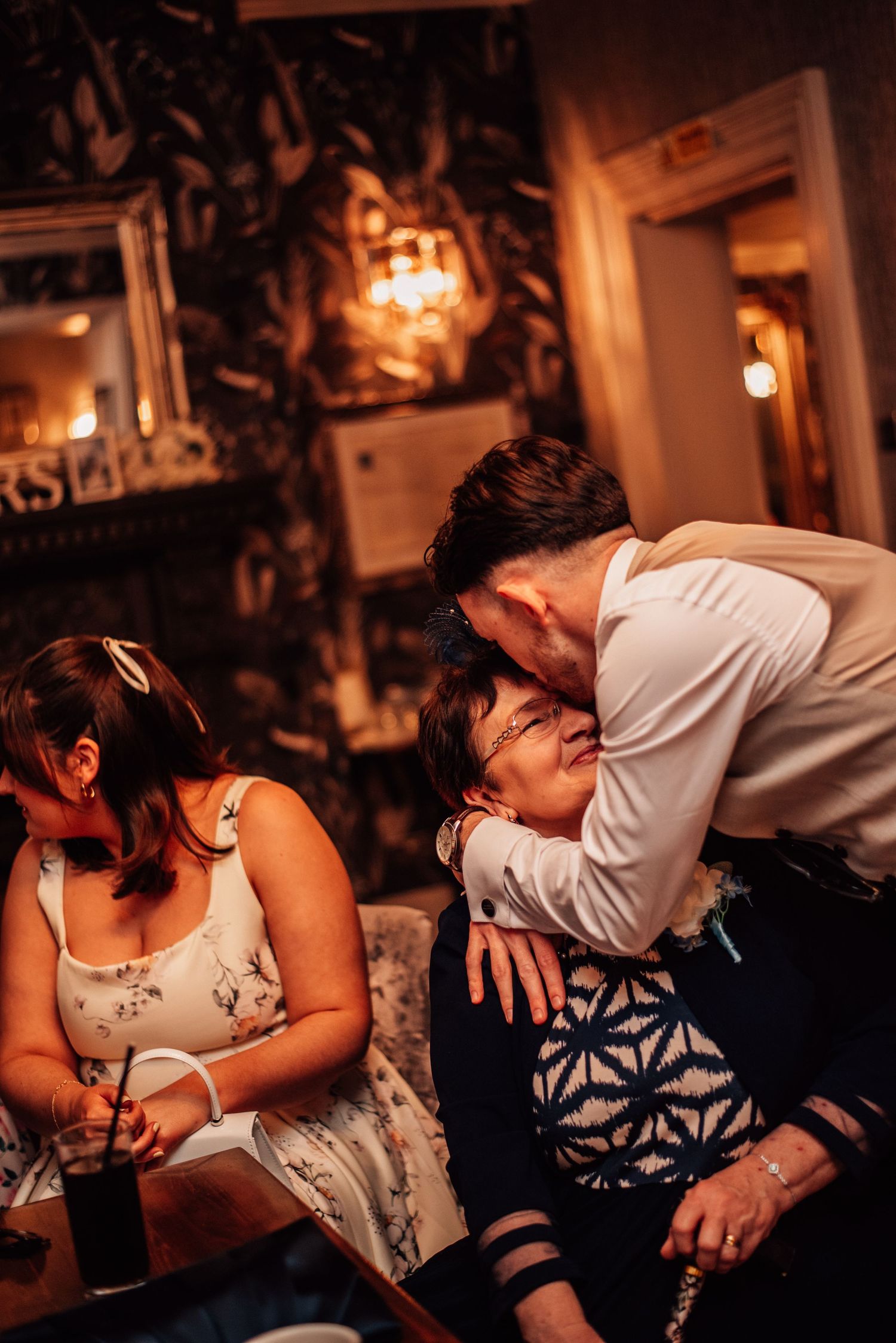 A man lovingly kisses an older woman on the cheek at a warmly lit restaurant gathering.
