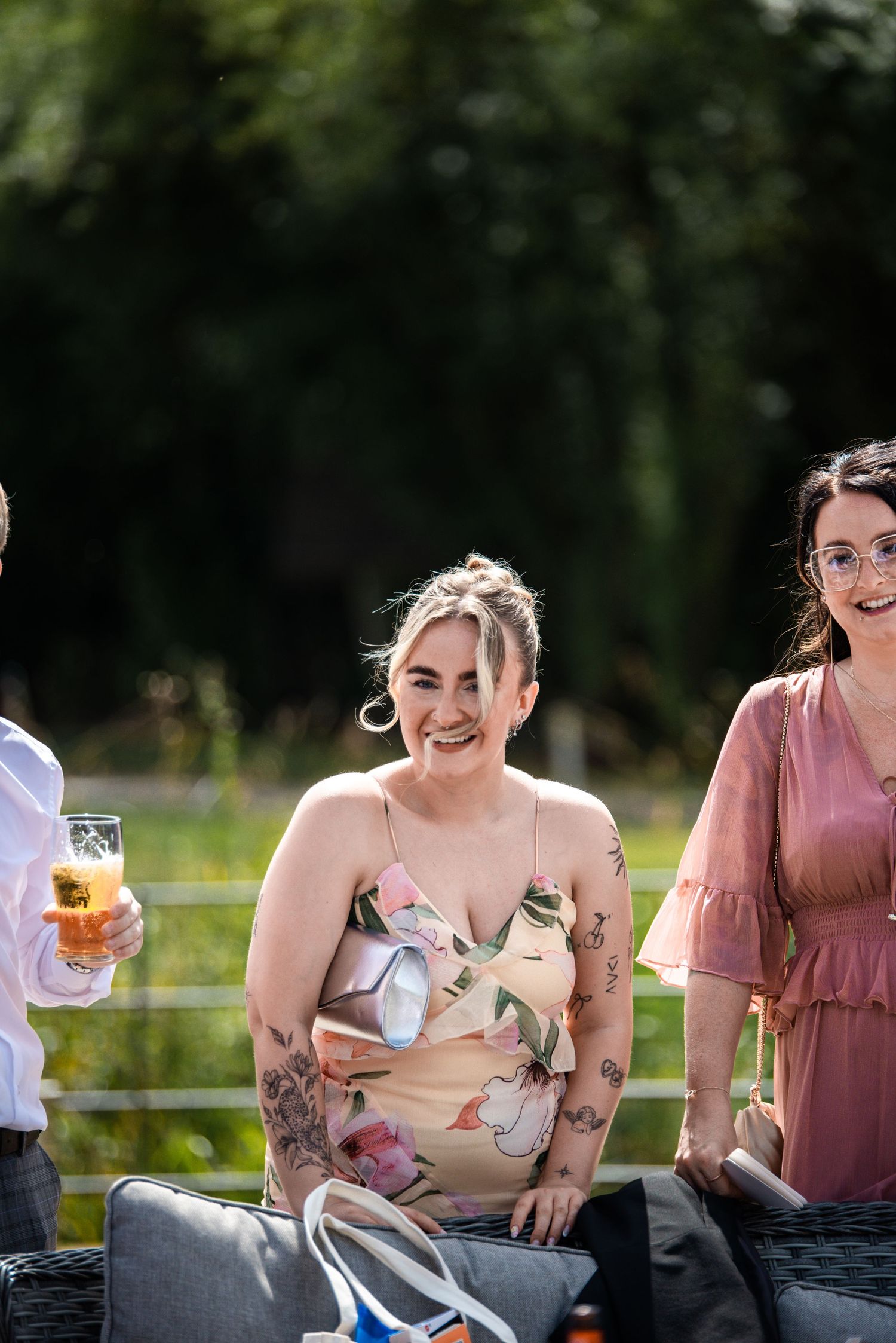 Women laughing and socializing outdoors at a summer garden party, wearing floral dress with tattoos.