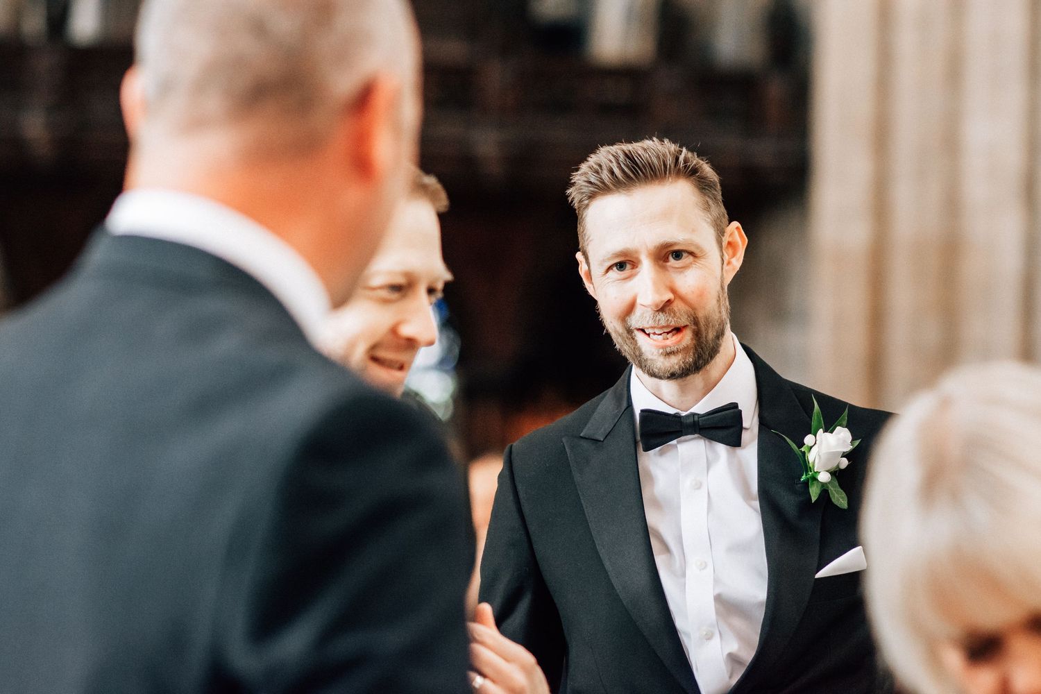 Groom in black tuxedo with bow tie and boutonniere smiling at wedding ceremony guests.