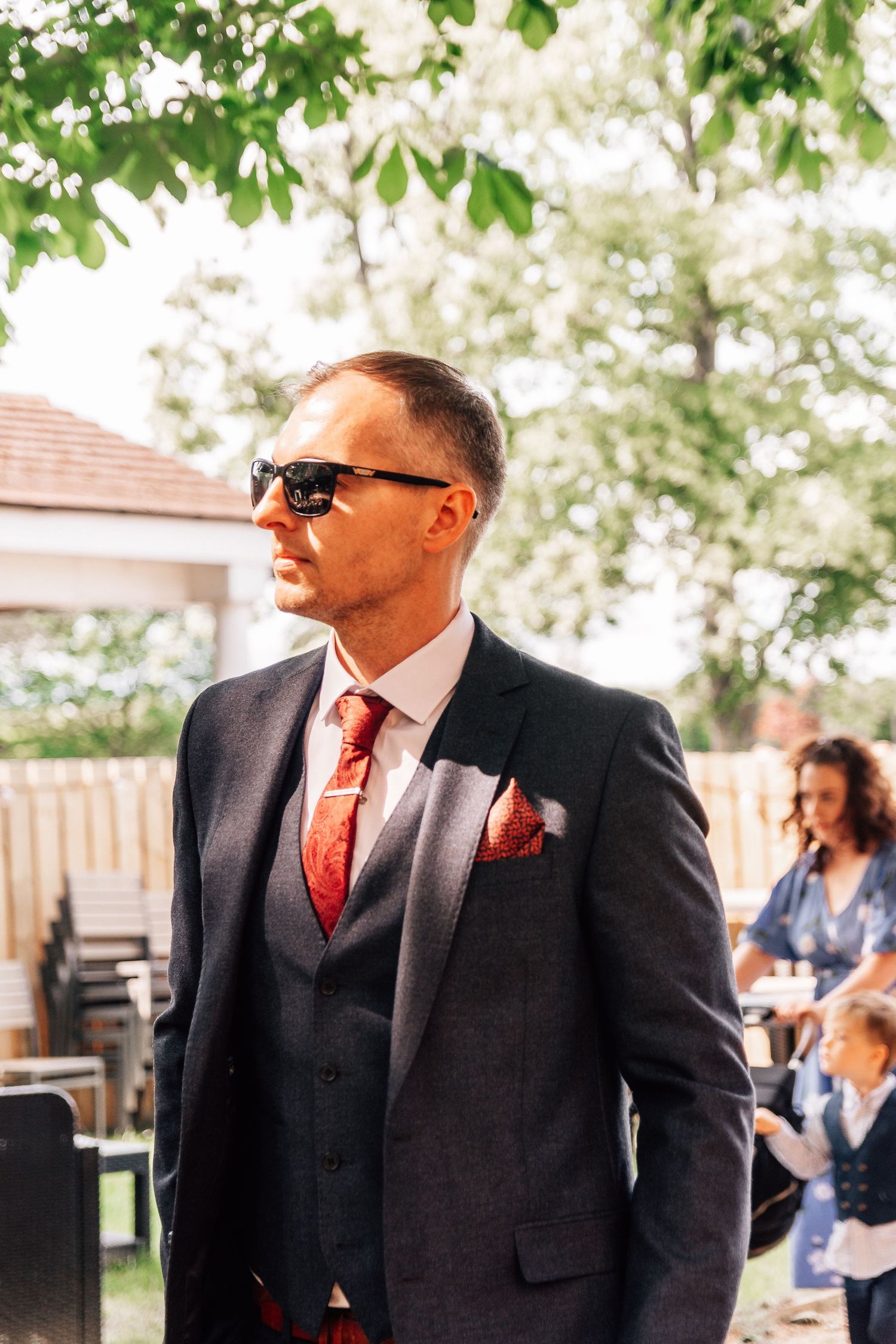 Stylish groom in dark suit with red tie and sunglasses at outdoor wedding.