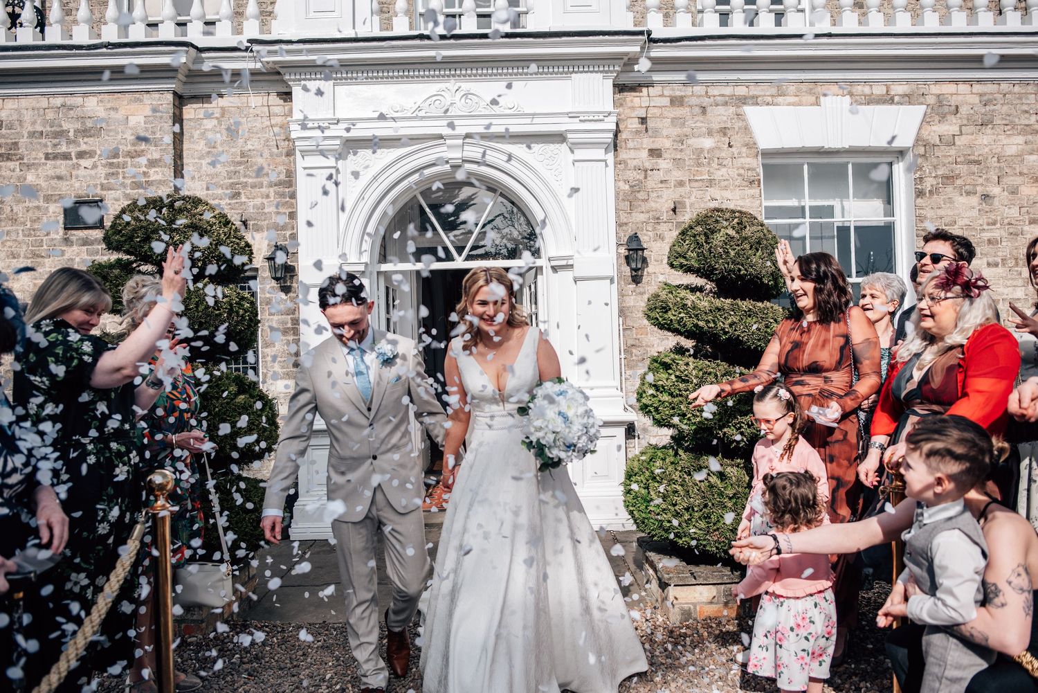 Newlyweds walk through confetti shower outside elegant manor house wedding venue with guests celebrating.