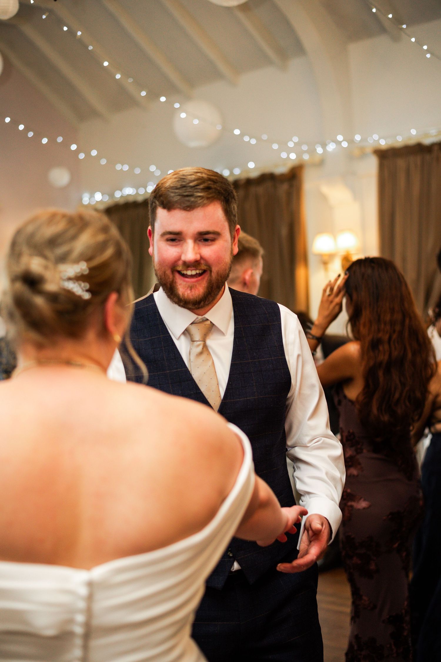 Smiling groom in navy waistcoat dancing with bride at wedding reception with fairy lights.