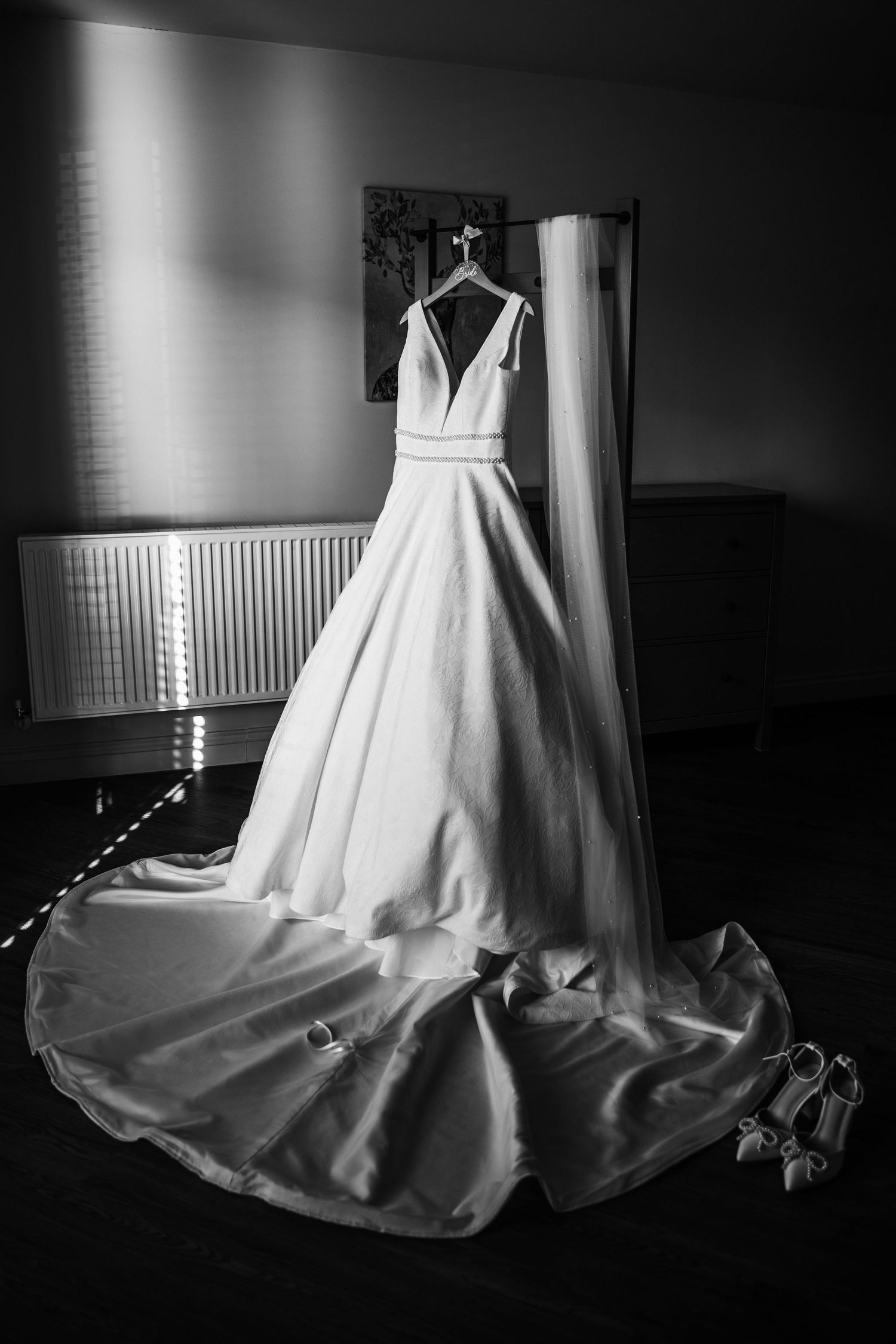 Black and white photo of an elegant bridal gown on a hanger with a long train and heels on the floor.