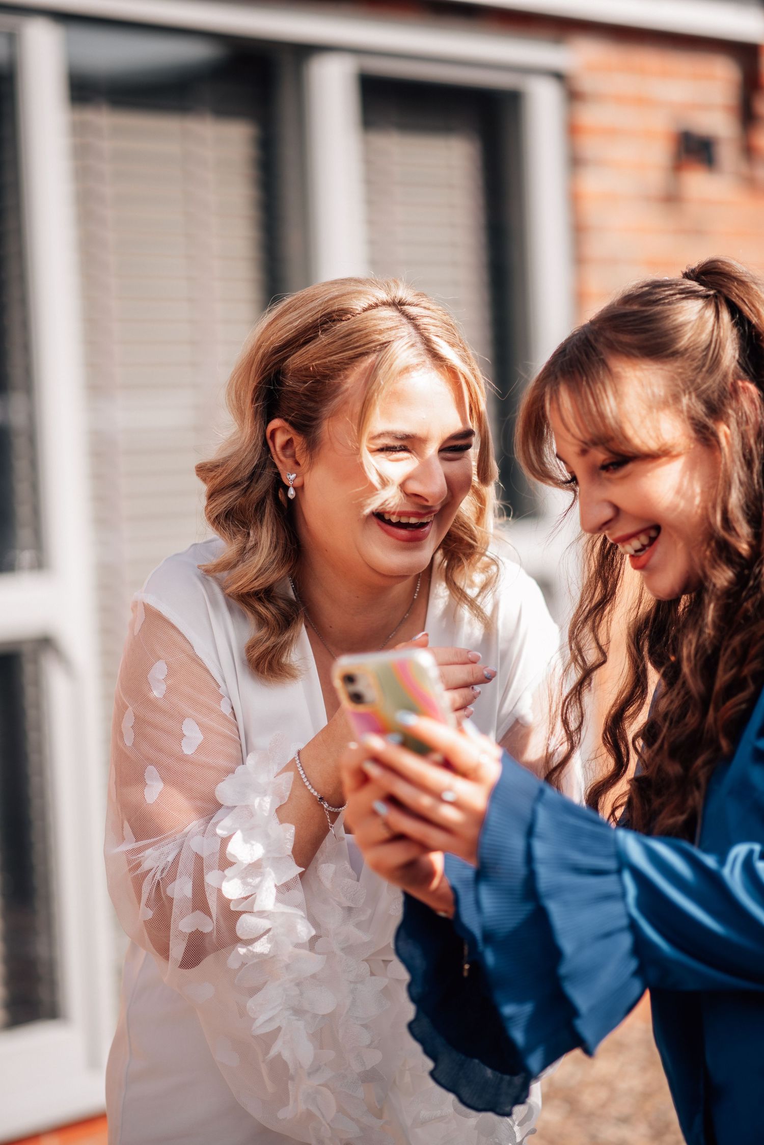 Bride and bridesmaid laughing together while looking at a smartphone outdoors.