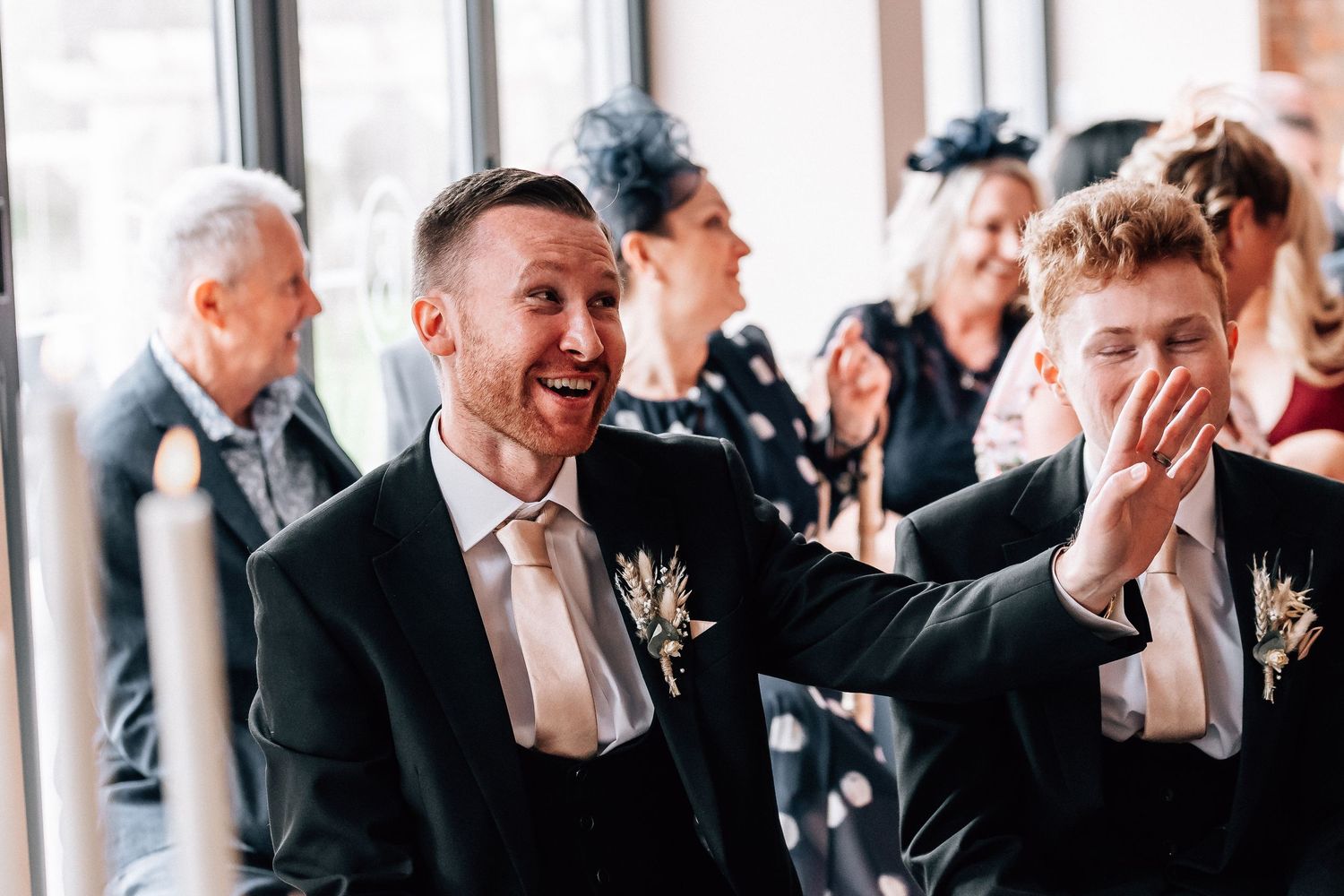 Two grooms laughing joyfully at their wedding ceremony, wearing dark suits with floral boutonnieres.