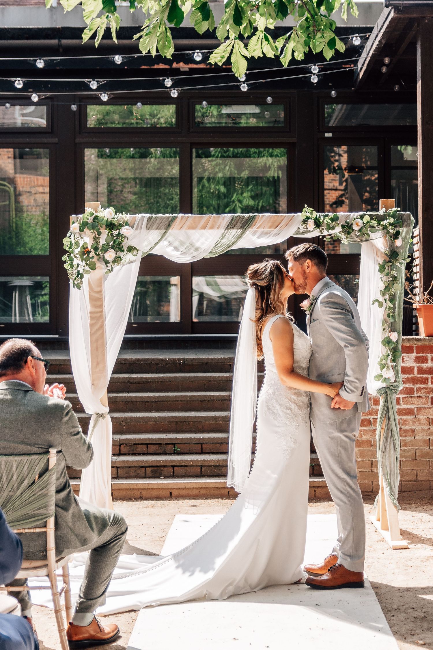 A romantic wedding couple shares a kiss behind flowing white curtains on a sunny patio.