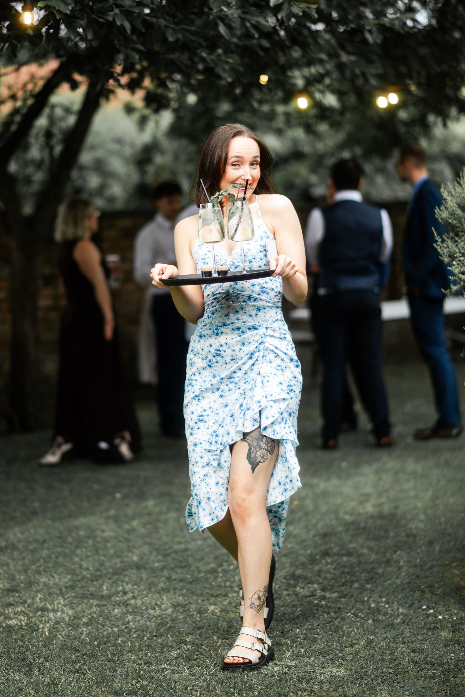 Woman in blue floral dress carrying a tray of drinks at an outdoor wedding reception.