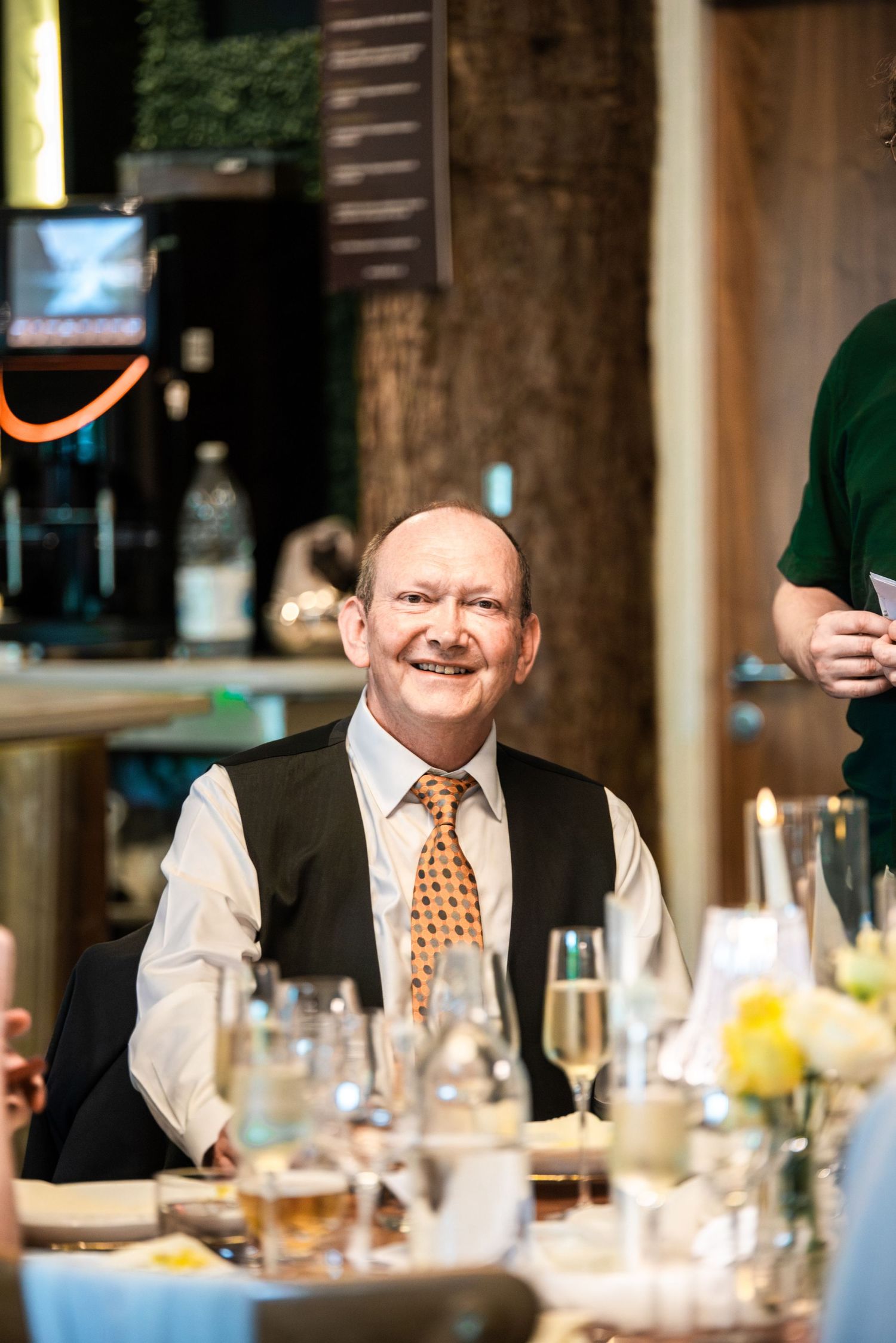 Smiling older gentleman in a vest and orange tie seated at an elegantly set wedding reception table.