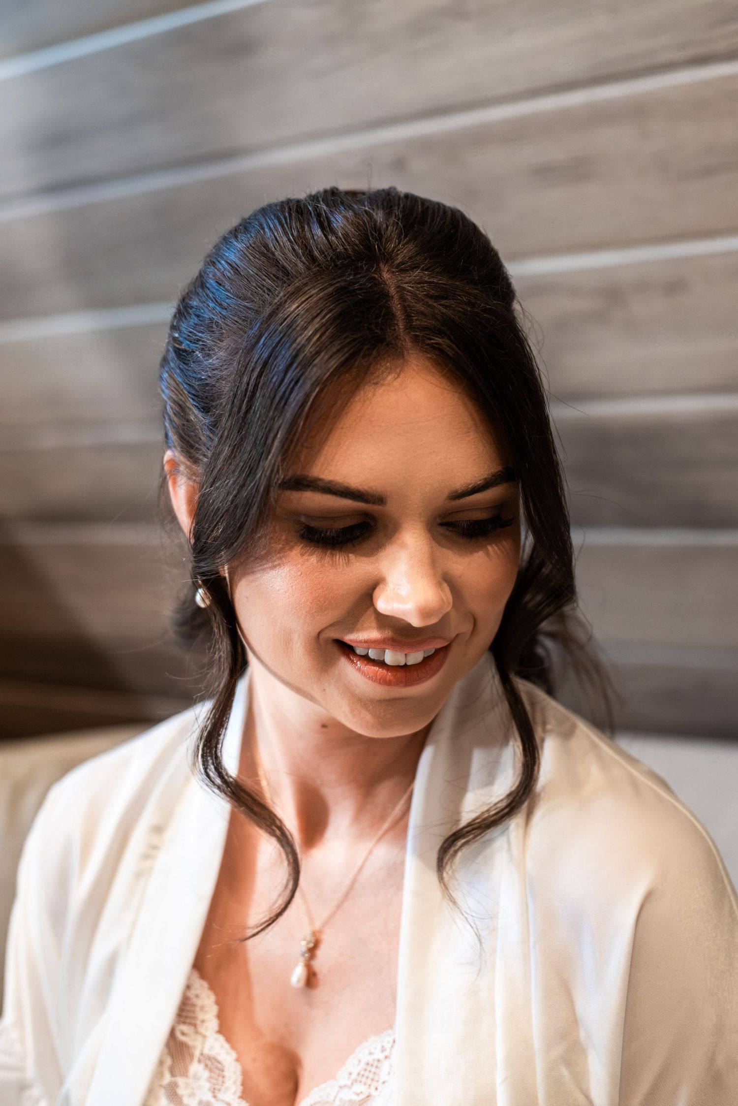 Smiling bride with dark updo hair, pearl earrings, and necklace wearing a white robe, looking down elegantly.