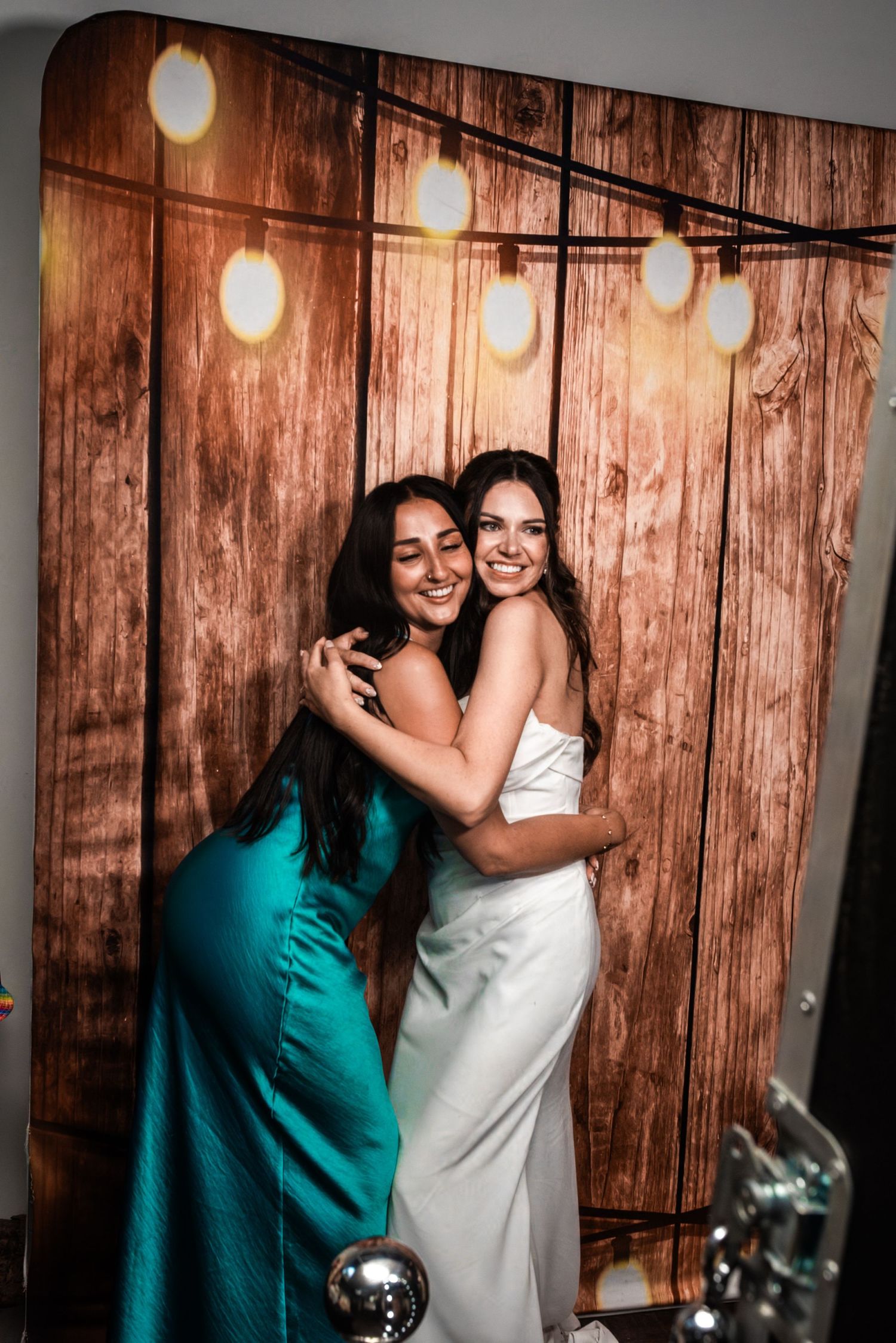 Two women hugging and smiling in front of a rustic wood backdrop with string lights at a wedding.