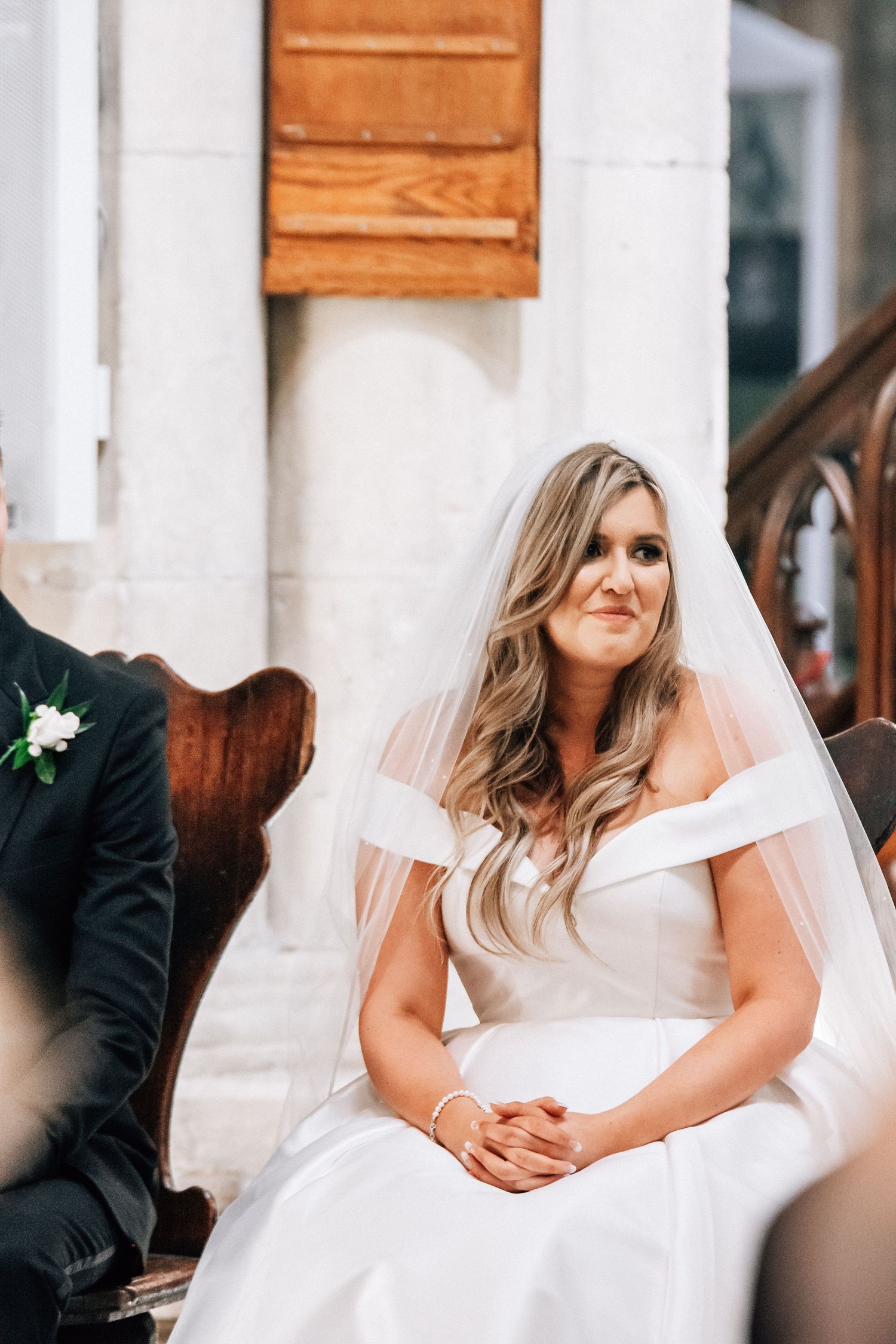 Bride in off-shoulder white gown and veil sits serenely during wedding ceremony in elegant church.