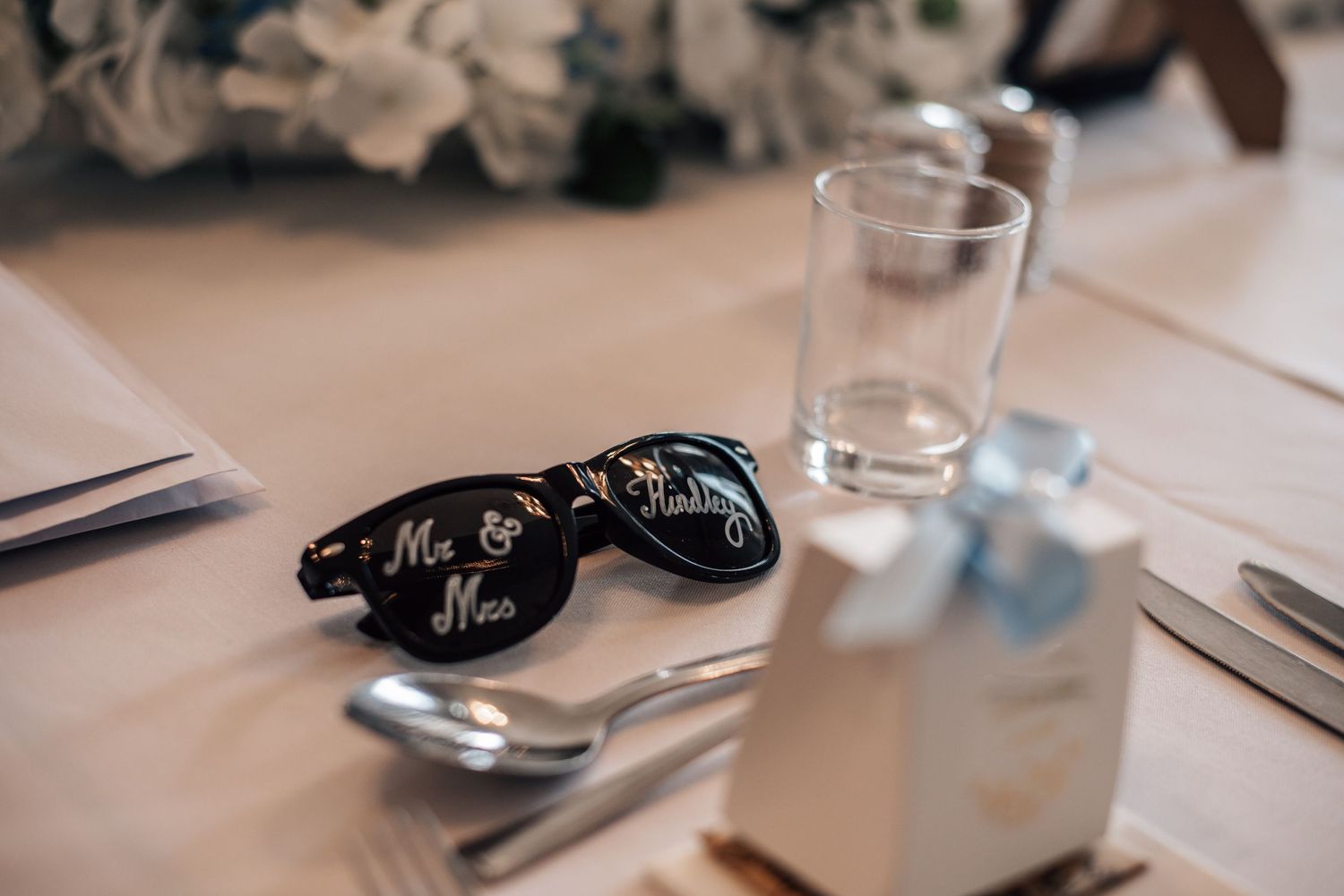 Wedding reception table with Mr. & Mrs. sunglasses, silverware, glassware, and a light blue gift box favor.