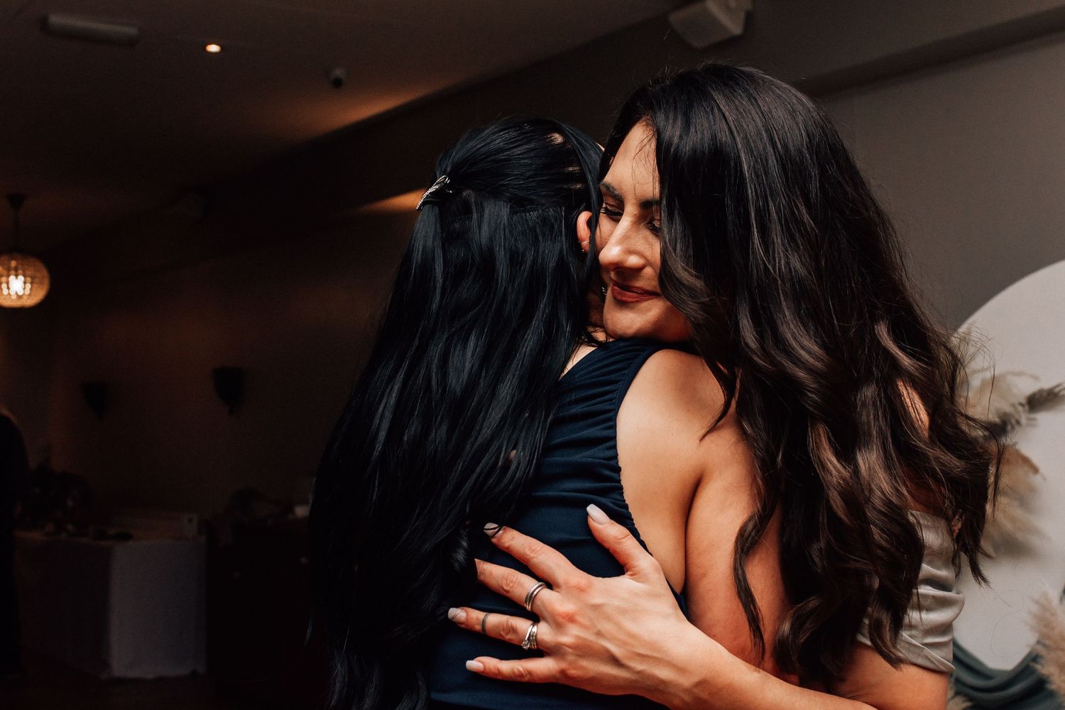 Two women share a warm embrace on a dance floor at an indoor event, both wearing elegant dark outfits.