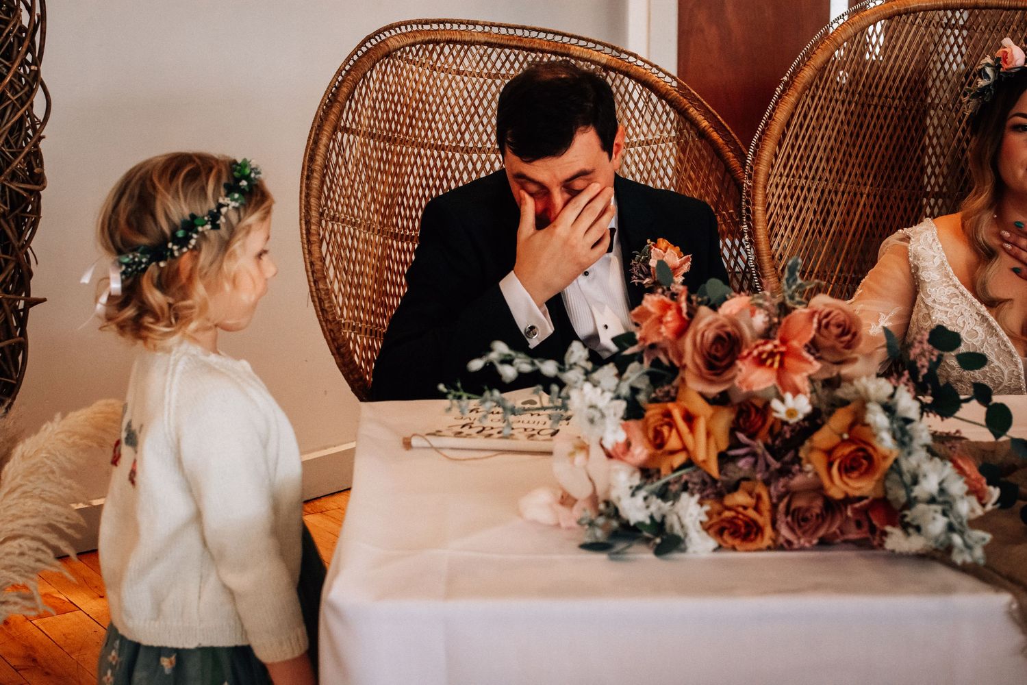 Emotional groom at table with floral arrangements as flower girl watches during wedding.