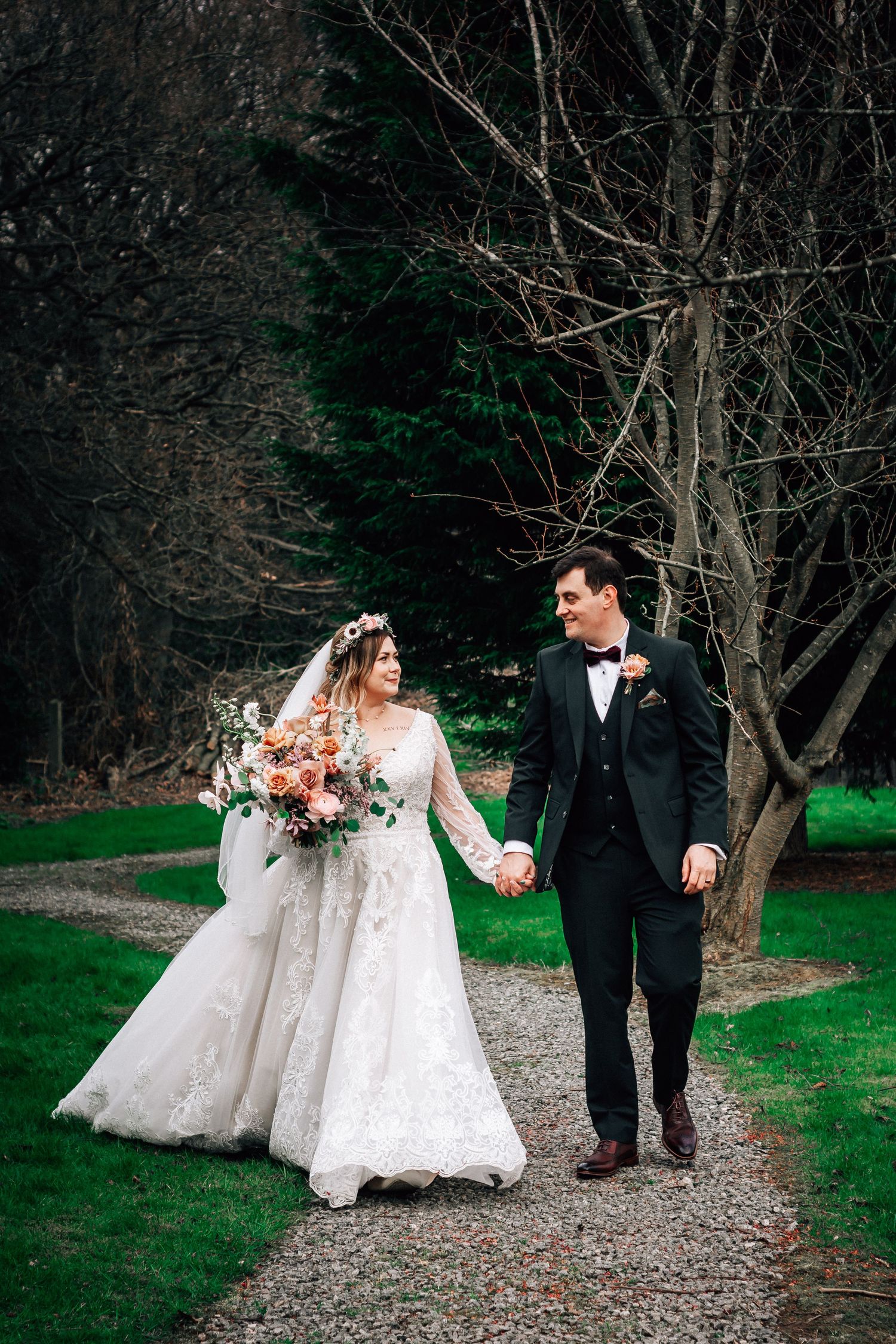 Bride and groom walk hand-in-hand on a garden path, she in lace gown with bouquet, he in dark tuxedo.