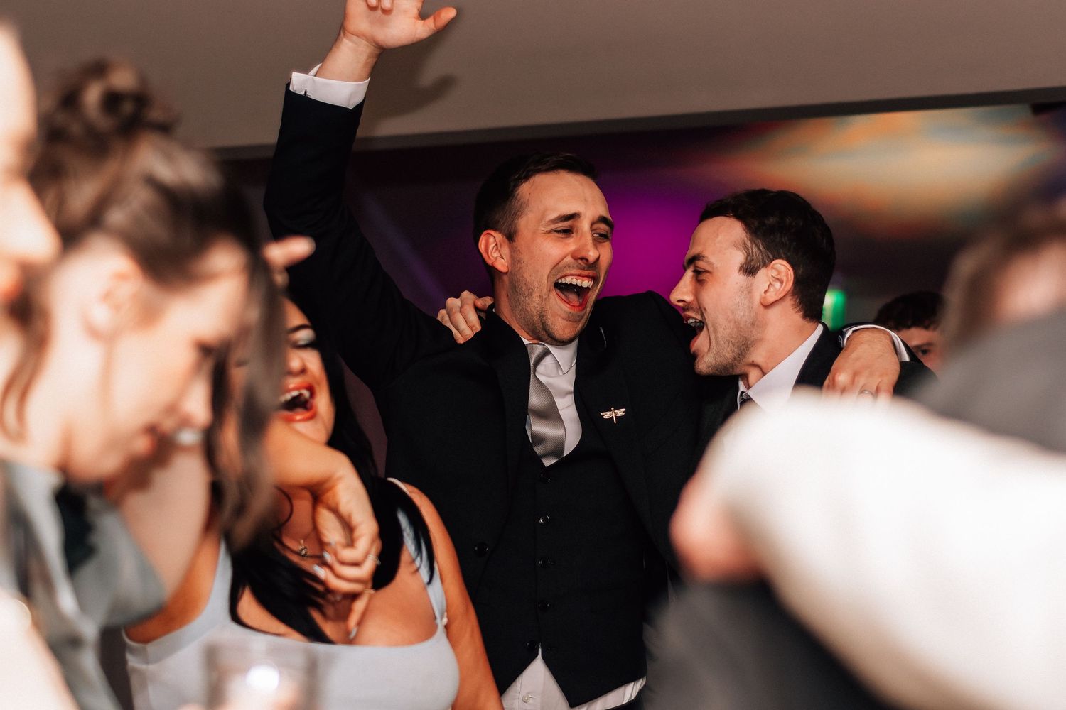 Joyful groom celebrating with guests on the wedding dance floor, laughing and raising his arm in excitement.