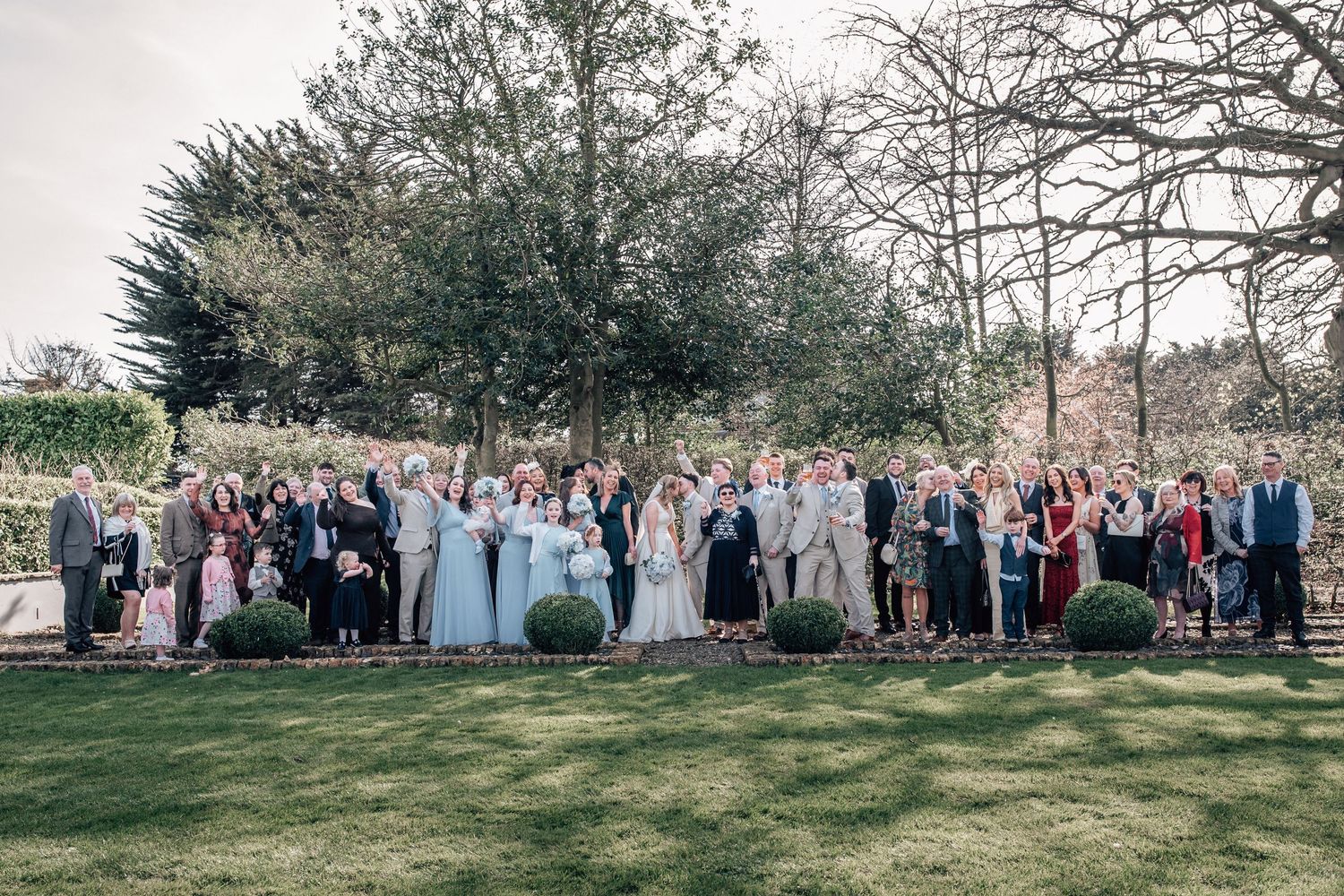 Large wedding group photo outdoors in a garden setting with bride, groom, and guests posed on a lawn.