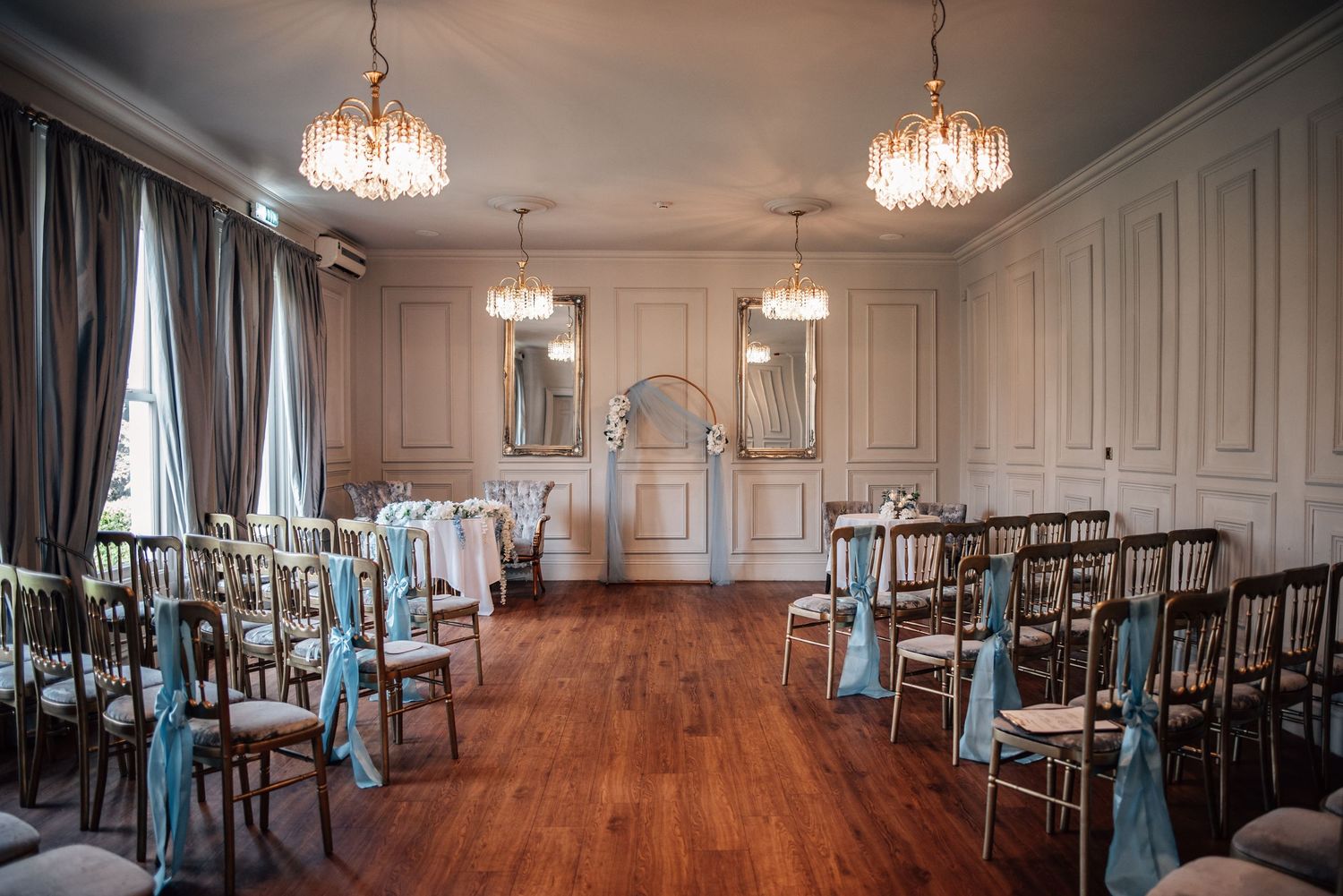 Elegant wedding ceremony room with crystal chandeliers, gold chairs with blue sashes, and a floral arch on hardwood floors.