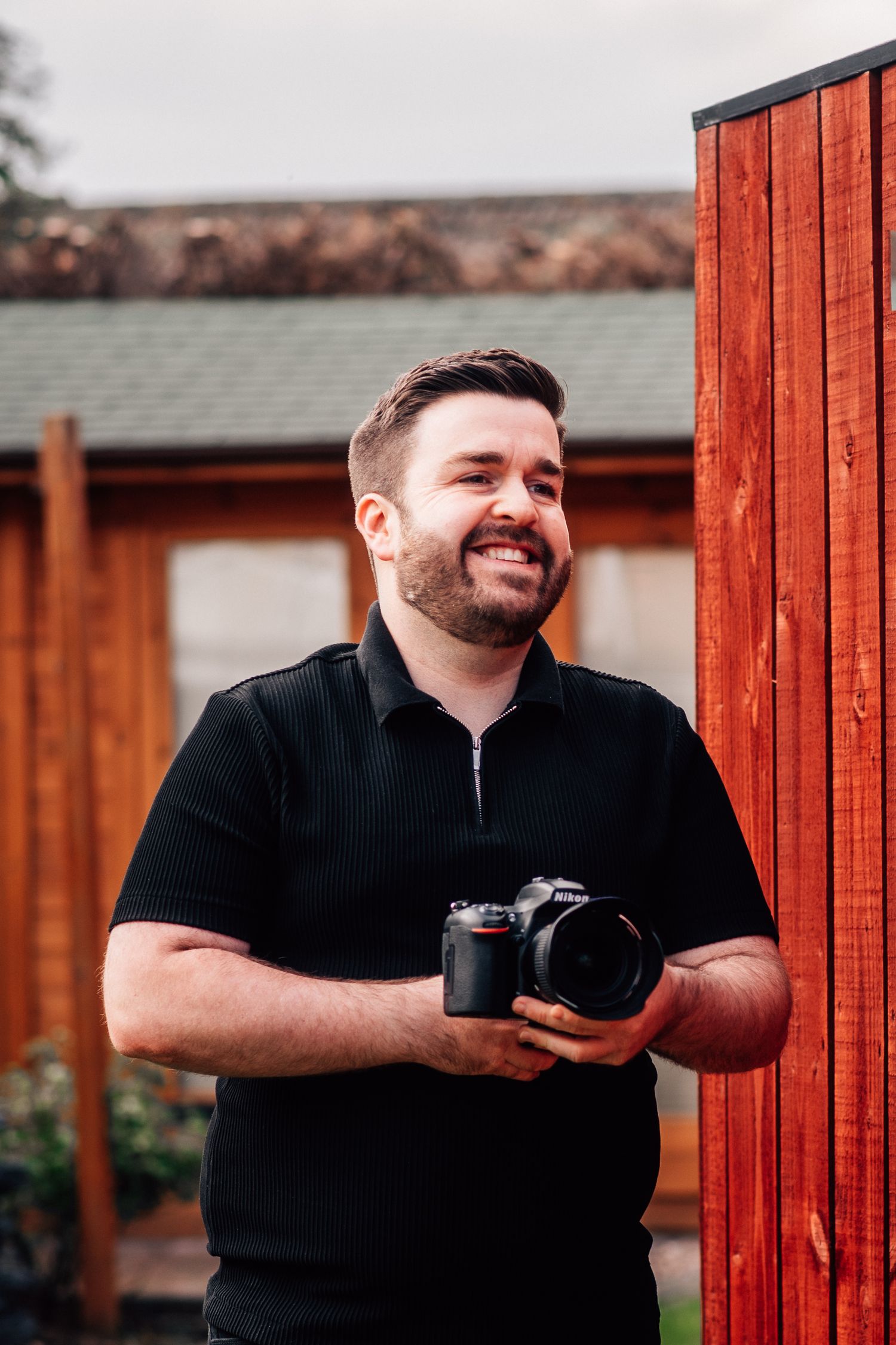 Smiling bearded man in black polo shirt holding a DSLR camera outdoors near a wooden fence.
