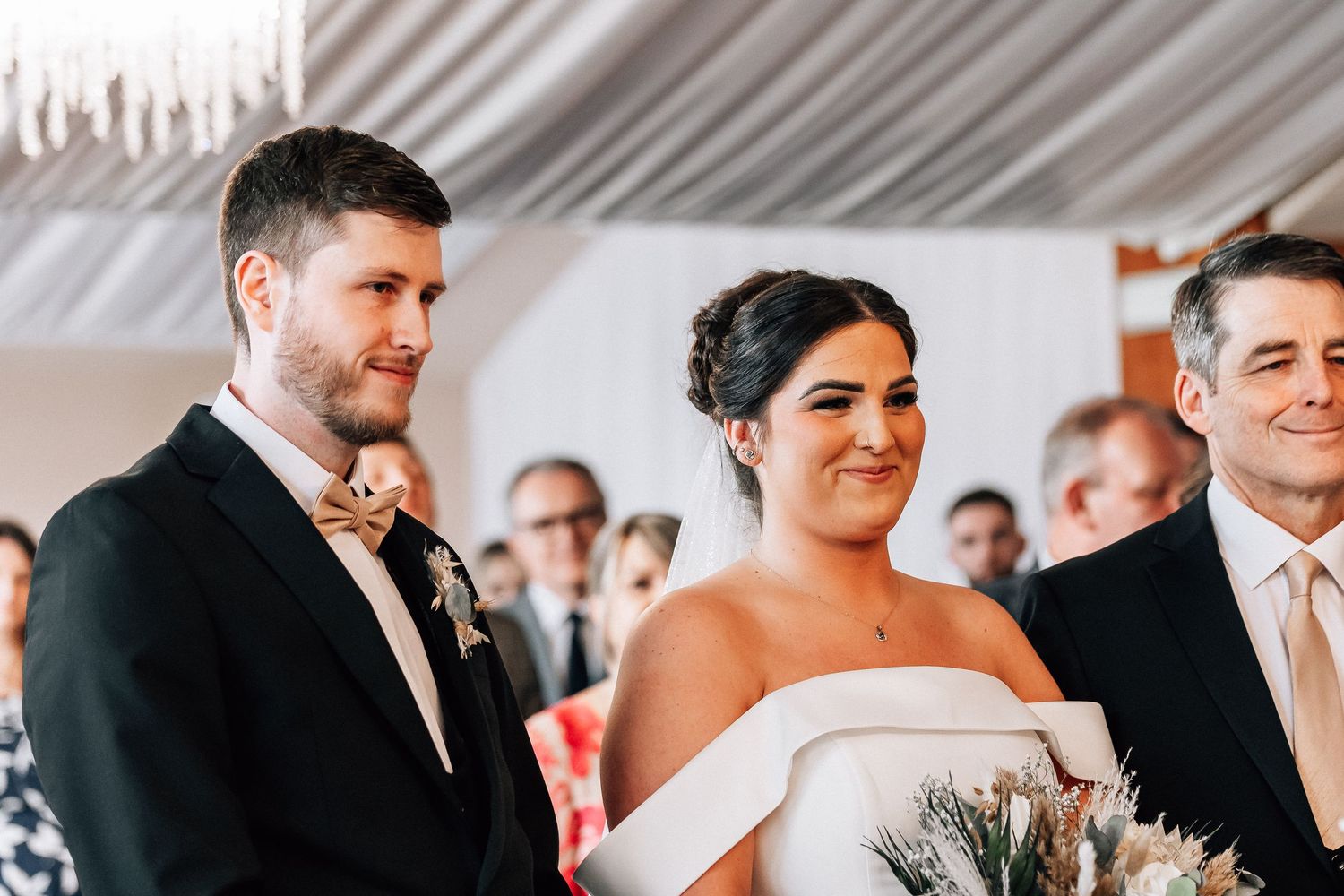 Bride and groom smiling during wedding ceremony, bride holding bouquet in elegant white gown, groom in black tuxedo.