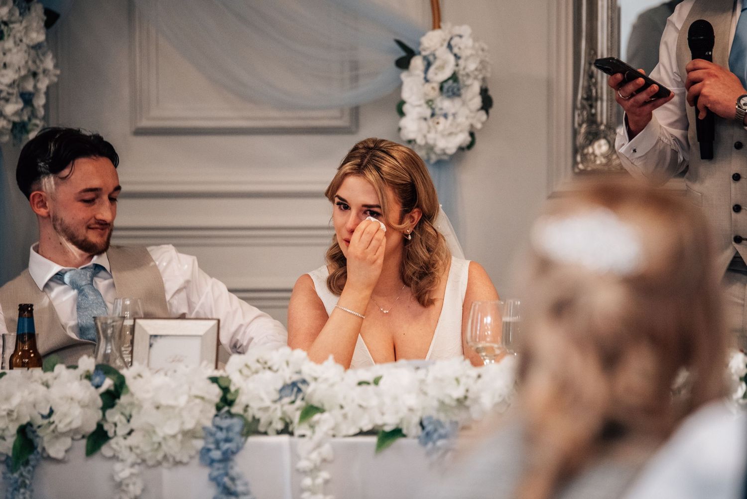 Emotional bride wiping tears during wedding reception speech, seated at floral-decorated head table with blue and white flowers.