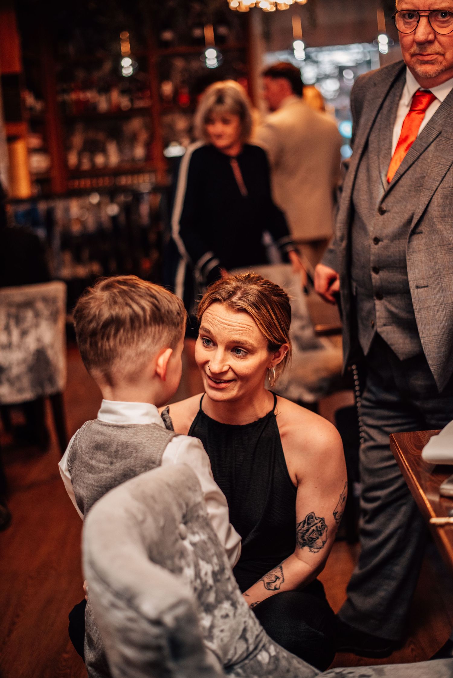 Woman in black dress with tattoo crouching to speak with young boy at wedding reception venue.