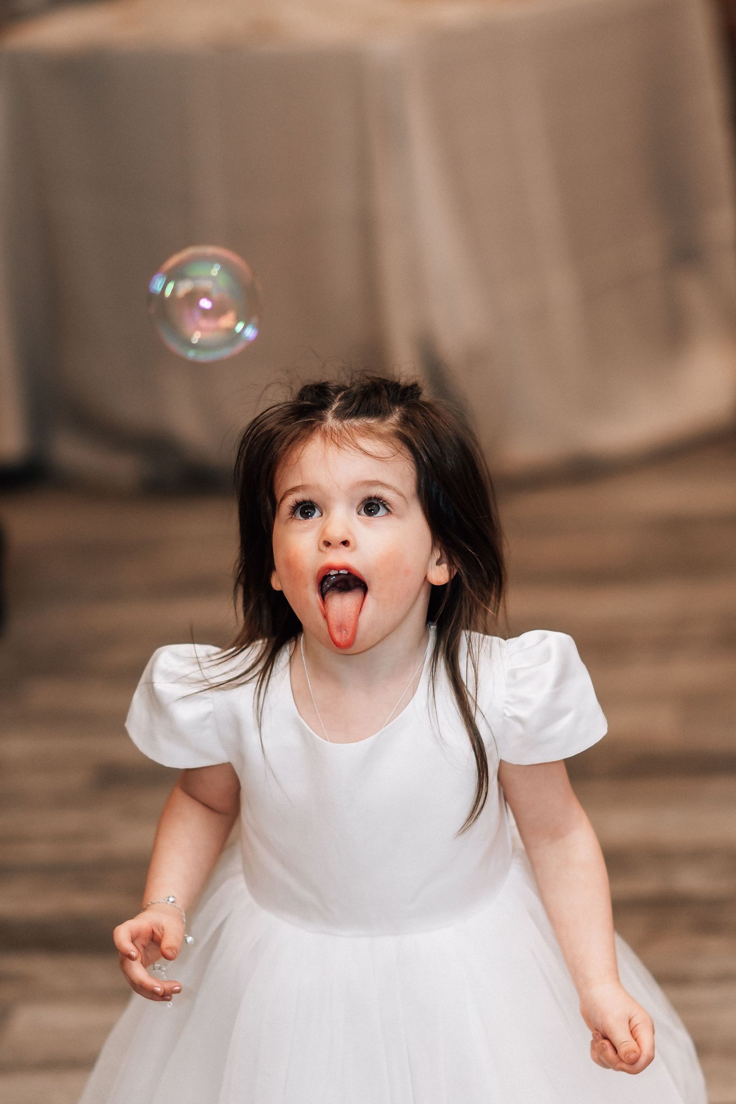 Young girl in white dress sticking out tongue trying to catch a soap bubble at an indoor event.