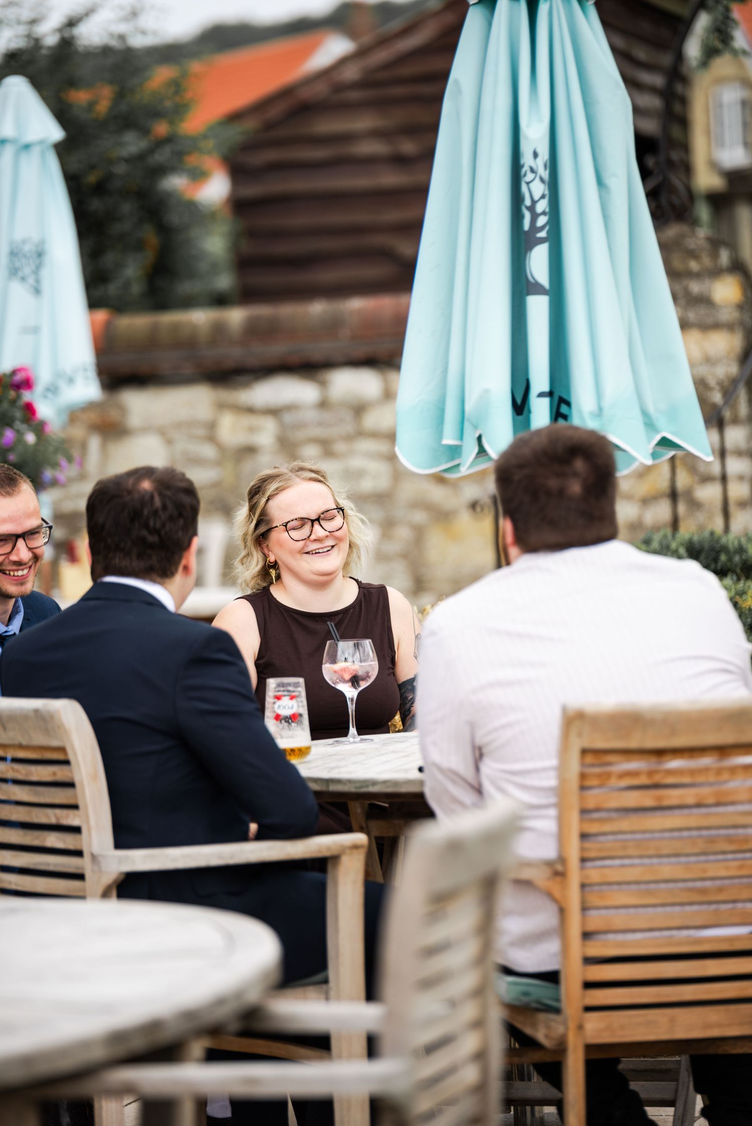 Friends enjoying drinks at an outdoor restaurant patio with teal umbrella and stone wall backdrop.