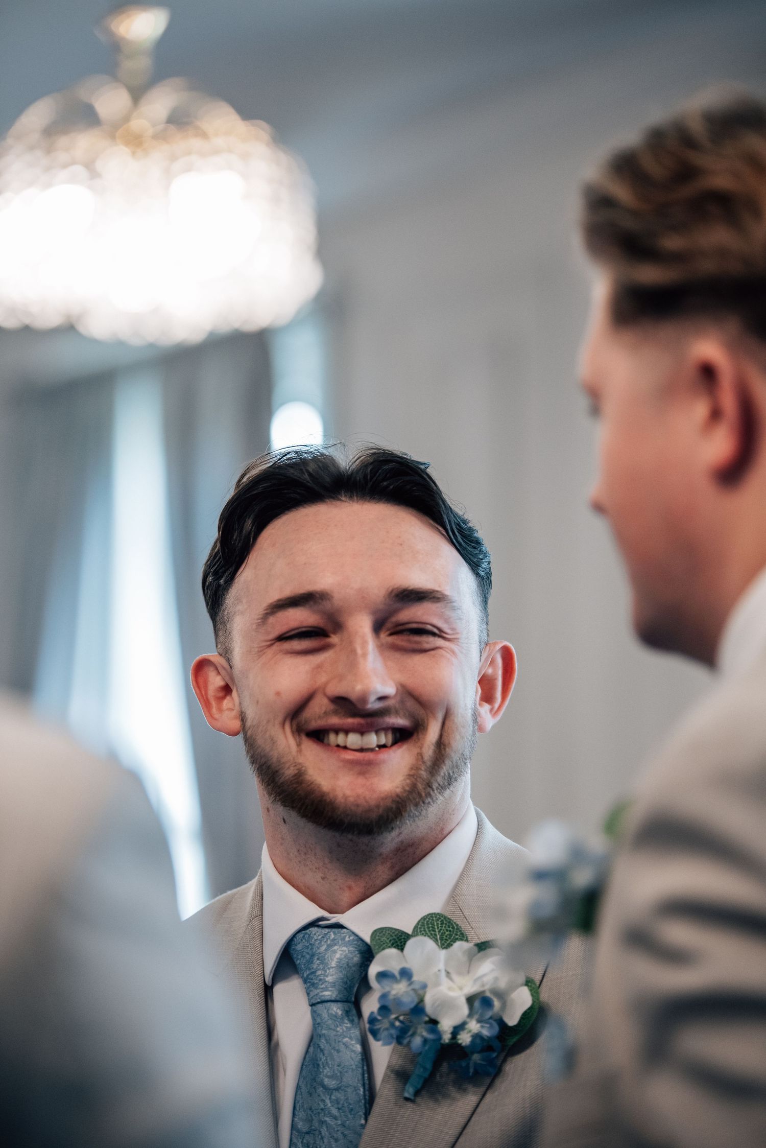 Happy groom in beige suit with blue tie and floral boutonniere smiling during wedding ceremony.