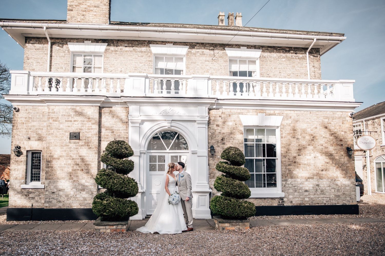 Bride and groom kissing outside an elegant brick manor house with topiary trees on their wedding day.