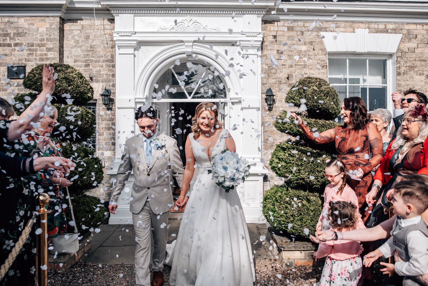 Bride and groom walk through confetti shower outside elegant venue surrounded by cheering wedding guests.